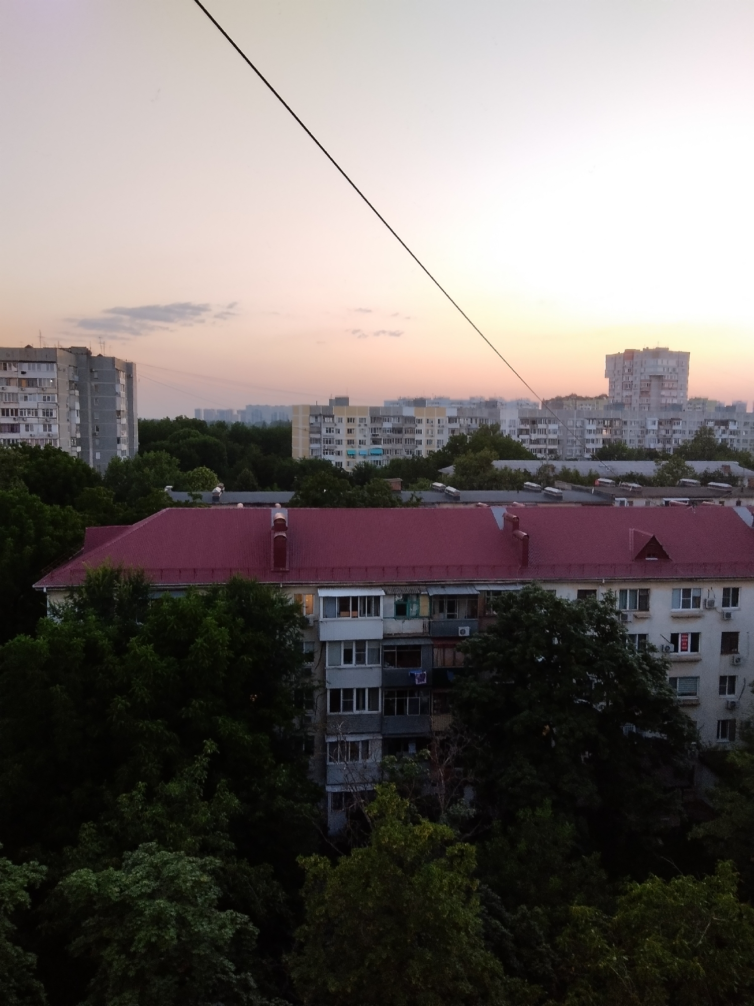 The image depicts a cityscape at dusk, likely in Krasnodar, Russia, characterized by a cluster of Soviet-era apartment buildings and dense foliage. In the foreground, a building with a prominent red-tiled roof is visible, flanked by lush green trees. This building has multiple stories with numerous windows and balconies, some of which are partially obscured by the trees. Beyond this structure, a panorama of residential high-rises stretches across the horizon. The sky above is a soft gradient of pale pink and orange, suggesting the setting sun, with a single, thin wire stretching diagonally across the upper portion of the frame. The overall impression is one of a residential district in the early evening.
