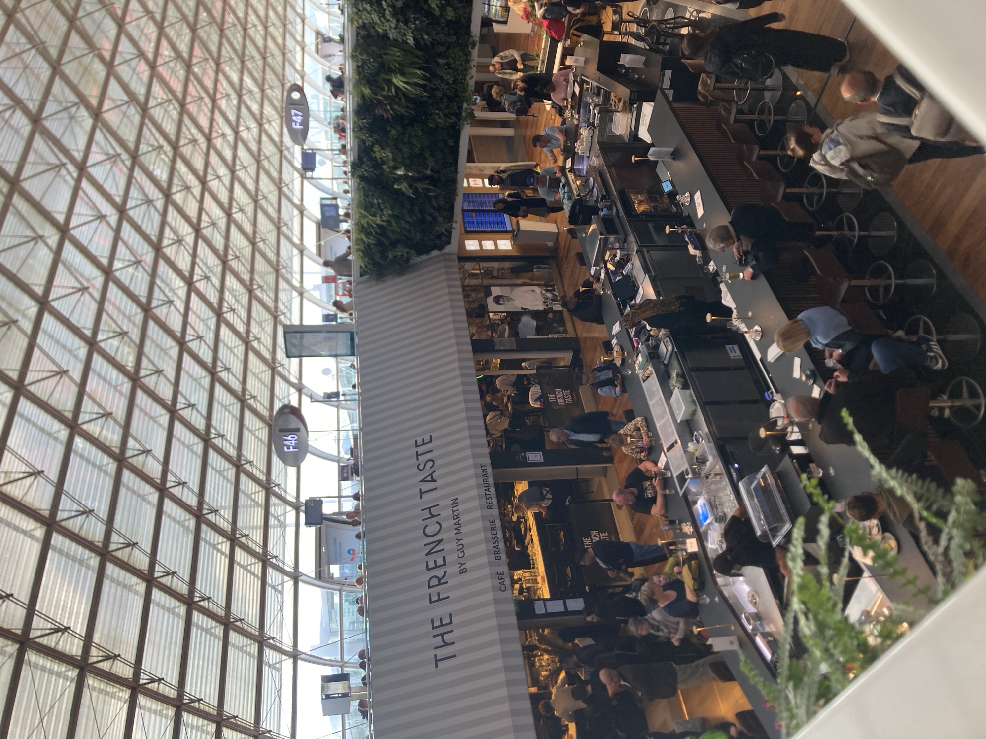 The image shows the interior of an airport restaurant called "The French Taste" by Guy Martin, located in Le Mesnil-Amelot, France. The restaurant features a long bar with stools where people are sitting, presumably eating and drinking. The background shows a large window with views of the airport's exterior, including gates labeled "F46" and "F47." The airport's architectural design includes a high ceiling with a visible grid-like structure. Green foliage decorates the space.