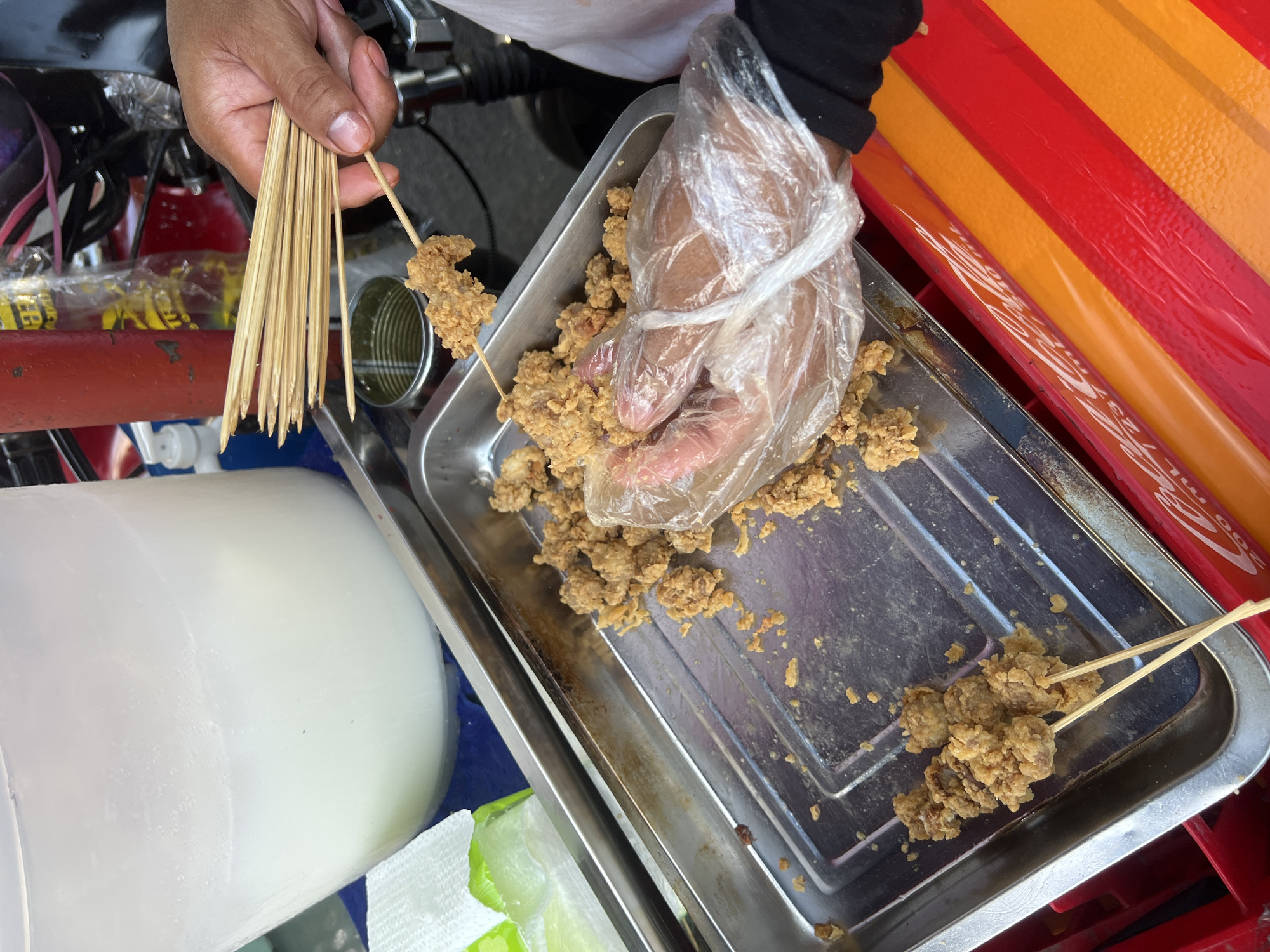 This image captures a close-up, overhead view of a street food vendor preparing deep-fried chicken bites on skewers. The food is served in a metallic tray. The scene appears to be in Davao City, Philippines, given the context of typical street food vendors in the area. A person's hand, wearing a clear plastic glove, is visible, holding a skewer with chicken bites. A stack of wooden skewers is also held by another hand, implying active preparation. In the background, a large white container likely holding a beverage, a red bottle, and parts of a vehicle suggest an outdoor setting. The overall impression is one of a bustling street-side food stall.