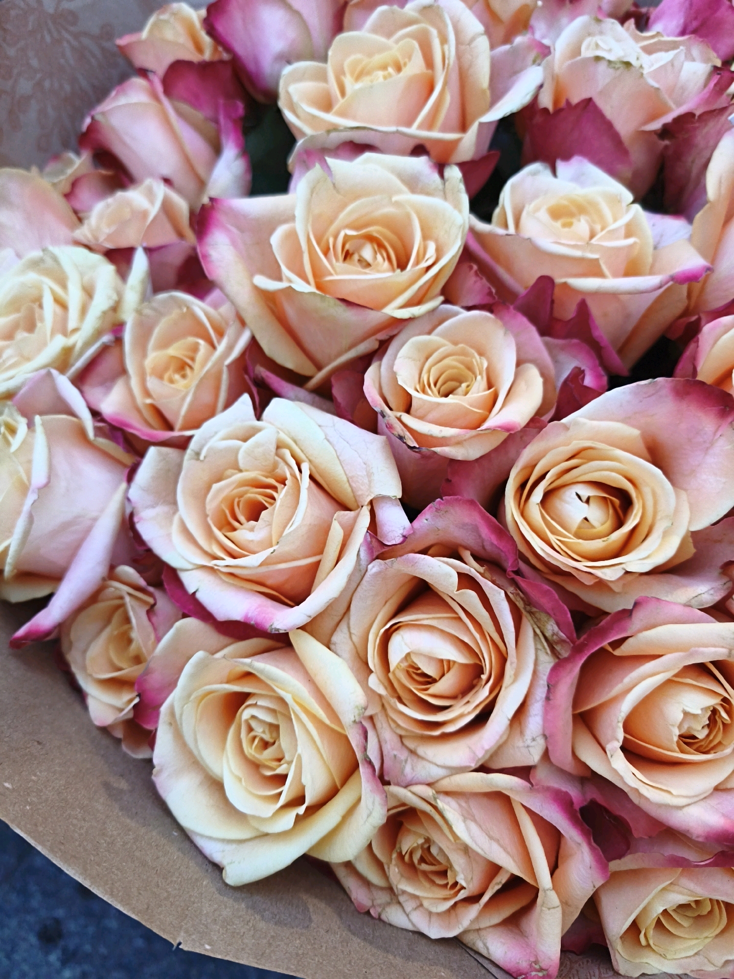 A close-up, top-down view shows a bouquet of peach and pink roses. The roses are tightly packed, with many layers of petals visible. Some of the roses have dark pink or fuchsia edges. The bouquet is wrapped in brown paper, which is slightly visible at the bottom of the image. The background is dark and out of focus. There is no indication of the city of Tallinn, Estonia, or any people.