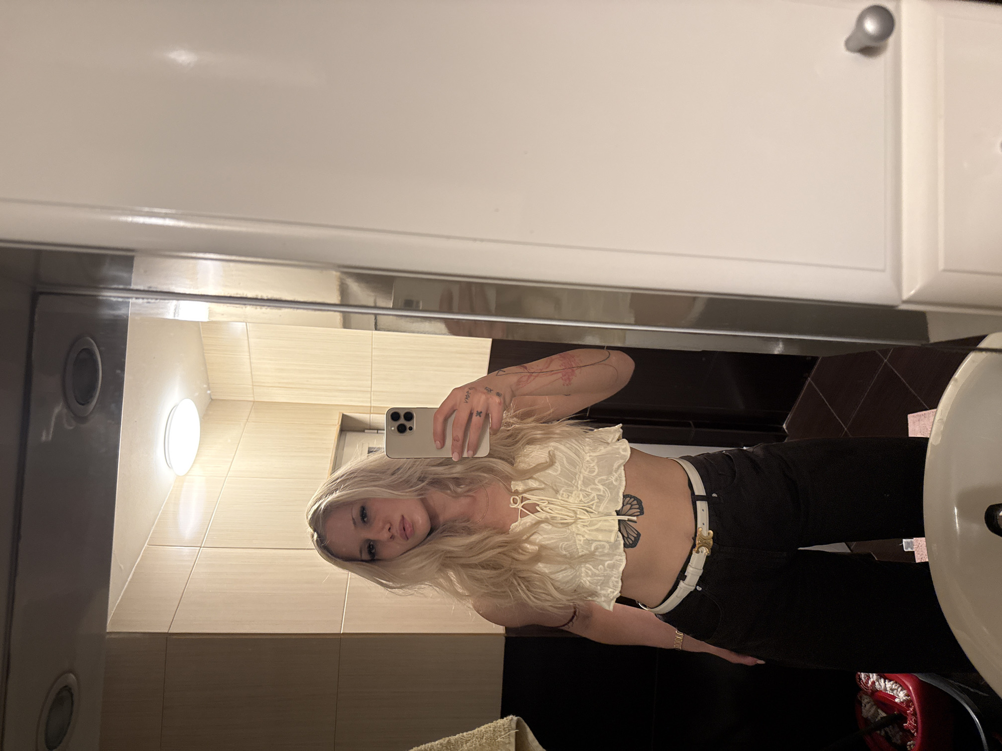 A young adult female is depicted taking a mirror selfie in a private bathroom, located in Sofia, Bulgaria. The subject has long, wavy blonde hair and is wearing a cream-colored, lace-up crop top with ruffles, black wide-leg trousers, and a white belt featuring a gold-toned "Celine" buckle. A dark butterfly tattoo is visible on her lower abdomen, an 'X' tattoo is on the back of her left hand, and multiple additional tattoos are present on both arms. She holds a light-colored smartphone with three camera lenses in her right hand, directed towards the mirror. The bathroom features a backdrop of light wood-grain patterned wall panels in the upper section, transitioning to dark brown/black square tiles below, which also form the flooring. A light-colored ceramic sink basin is partially visible in the lower foreground. The ceiling contains two recessed circular lights and one central circular flush-mount light fixture. A yellow towel hangs on a rail to the left, and a white cabinet door with a silver knob is visible on the far right. A red bucket is situated on the floor to the left of the subject.