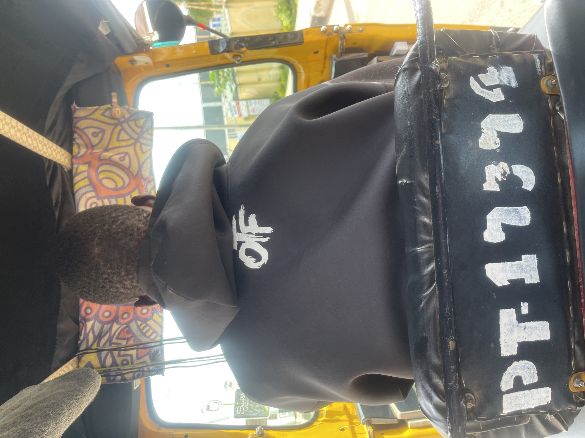 An individual is seated in the driver's seat of a yellow auto-rickshaw, their back to the camera. They are wearing a black hooded sweatshirt with the white letters "OTF" emblazoned on the back. The auto-rickshaw's interior is visible, including a colorful, patterned fabric hanging from the roof and a black seat with white lettering on the back that appears to read "PT-13374". The scene is outdoors, with a building and trees visible through the open window of the auto-rickshaw, suggesting a location in a city like Gura Top, Nigeria. The lighting indicates it is daytime, likely with bright sunlight.