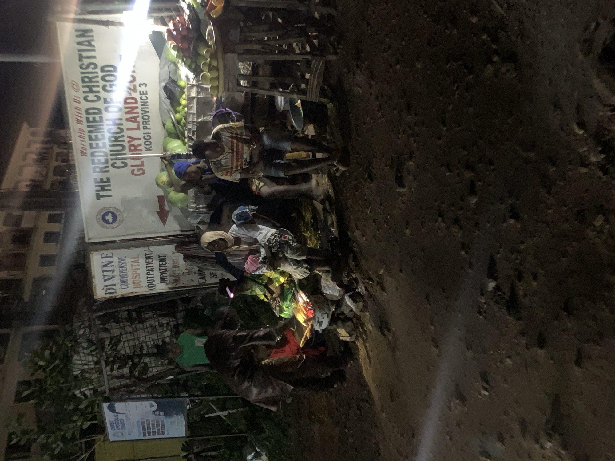 The image depicts an outdoor scene at night, likely in the city of Lokoja, Nigeria, given the presence of signage related to "Kogi Province." In the foreground, an individual is tending to a small cooking fire made of stones and embers, with what appear to be corn cobs roasting over it. This person is wearing dark clothing and is bent over the fire. To the right of the fire, three people are seated. One, wearing a white headscarf and a light-colored garment, is looking towards the fire. Another, wearing a striped t-shirt and denim shorts, is looking down. The third person, behind the seated individuals, is partially visible and appears to be wearing a blue head covering and a green patterned outfit.
Behind the seated individuals is a stall laden with fruits and vegetables, including green melons, apples, and what look like tomatoes or peppers. The stall is supported by rustic wooden structures. Further in the background, a large banner is prominently displayed, reading "Worship With Us @ THE REDEEMED CHRISTIAN CHURCH OF GOD GLORY LAND-ZONE... KOGI PROVINCE 3." To the left of this banner, there are other signs, one of which displays a picture of a man and text that includes "CHRIST APOSTOLIC CHURCH." The overall environment appears to be a somewhat unpaved area with a dirt ground surface, and a building or structure is visible in the far background, illuminated by lights. The scene suggests a street-side market or food stall operation occurring after dark.