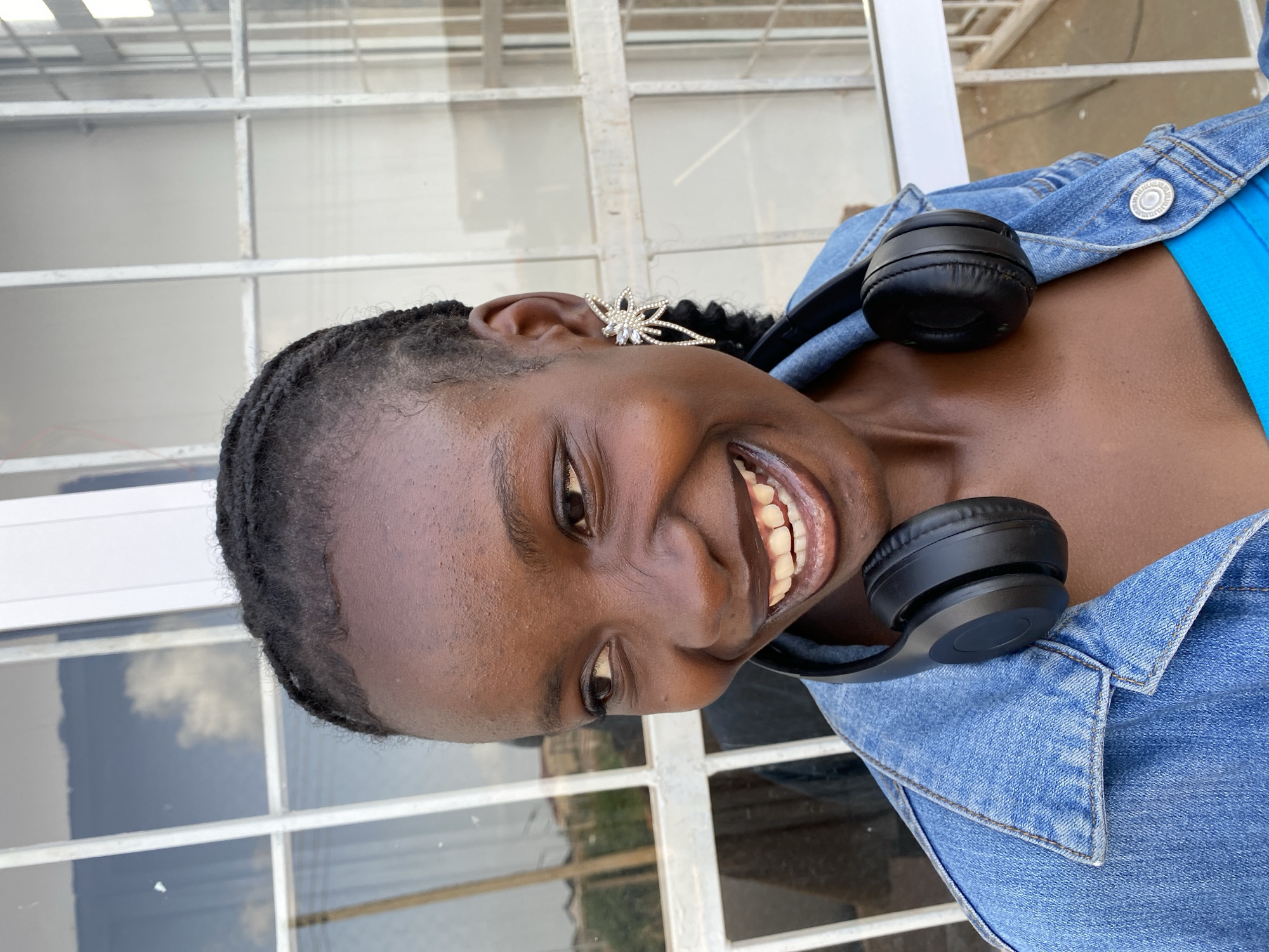 This media file is a portrait of a young Black woman. She is looking directly at the camera with a wide smile, revealing her teeth. Her hair is styled in braids, and she is wearing a large, sparkly earring in the shape of a starburst with crystals. She has on a blue denim jacket over a bright blue top. A pair of black headphones rests around her neck, with one earcup visible near her shoulder.
The background appears to be an outdoor setting with a white-framed structure, possibly windows or a fence, with reflections of clouds and greenery visible. The lighting suggests it is daytime. The image is taken from a close-up perspective, focusing on the woman's face and upper body. Her expression conveys joy and friendliness. There is no visible text in the image. The setting provides hints of being in Bar Jirgi Summa, Nigeria.