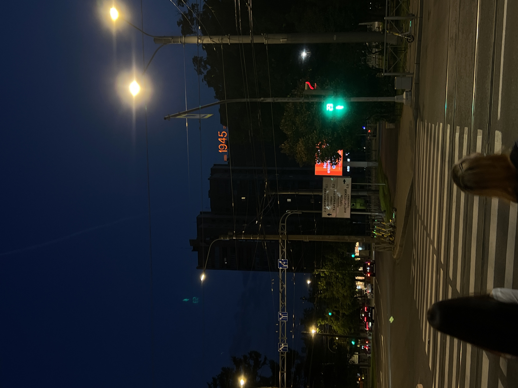 The image captures an evening street scene in Saint Petersburg, Russia, likely near the intersection of Prospekt Mira and ulitsa Karbysheva, given the signage visible. A crosswalk dominates the foreground with two individuals, one with dark hair and the other with blonde hair, standing on it, their backs to the camera. Behind them, a wide street with multiple lanes is illuminated by streetlights. Several cars, including red and white vehicles, are driving on the road. A traffic light displays a green signal for pedestrians, with the number "29" visible within the pedestrian icon. To the right, a series of yellow electric scooters are parked alongside a metal fence. Further down the street, a large building stands tall, its facade illuminated by the city lights. Notably, the building displays a large orange digital sign reading "1945" and another sign with Cyrillic text. Trees line the sidewalks, their foliage visible against the dark blue night sky. The overall atmosphere is that of a typical urban setting at dusk.