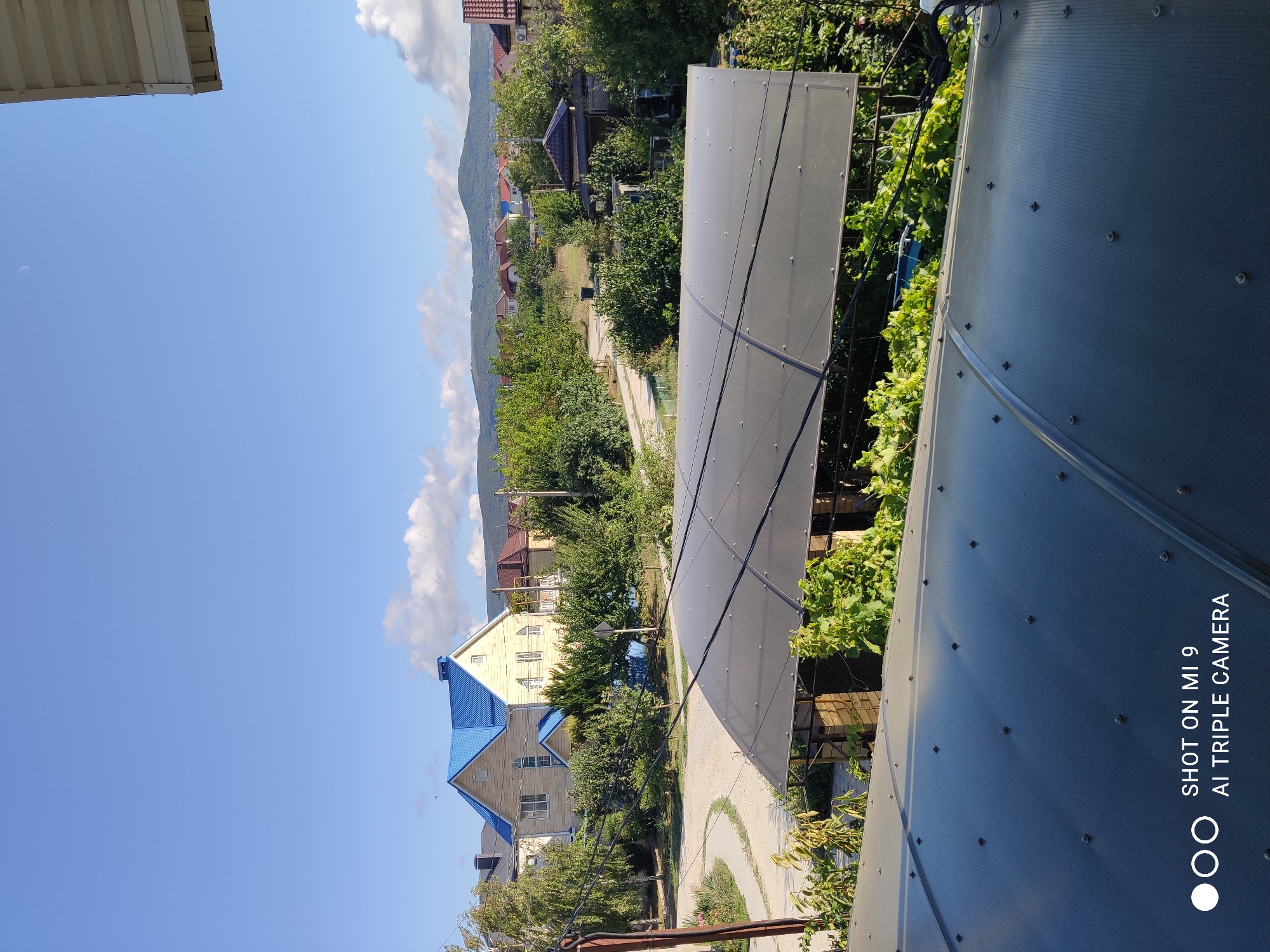 The image captures an outdoor scene, likely in Anapskaya, Russia, during the daytime. The viewpoint is elevated, looking over a residential area with several houses, lush green trees, and rolling hills in the background. A prominent feature in the foreground is a dark, translucent, curved roof structure, possibly a greenhouse or a covered patio. Beyond this, a beige-colored two-story house with a bright blue roof stands out. A dirt road winds through the property. The sky is a clear blue with scattered white clouds. The overall impression is one of a peaceful, rural or suburban environment.