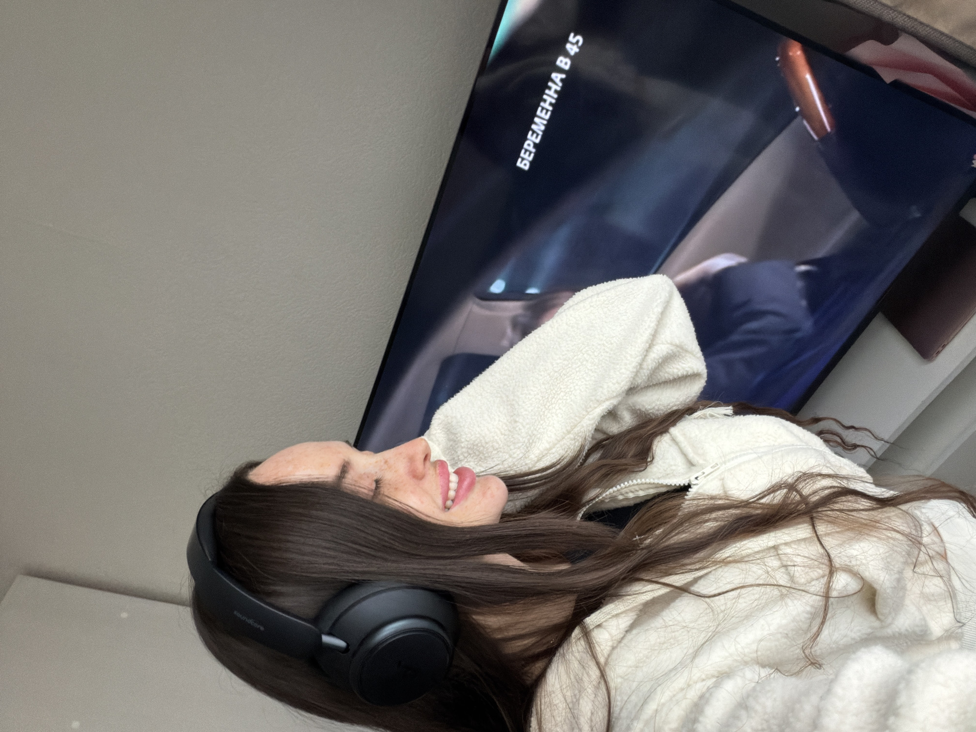 A person wearing headphones and a white jacket is lying down, watching a TV screen. Text in Russian is visible on the screen. The location cannot be determined from the image.