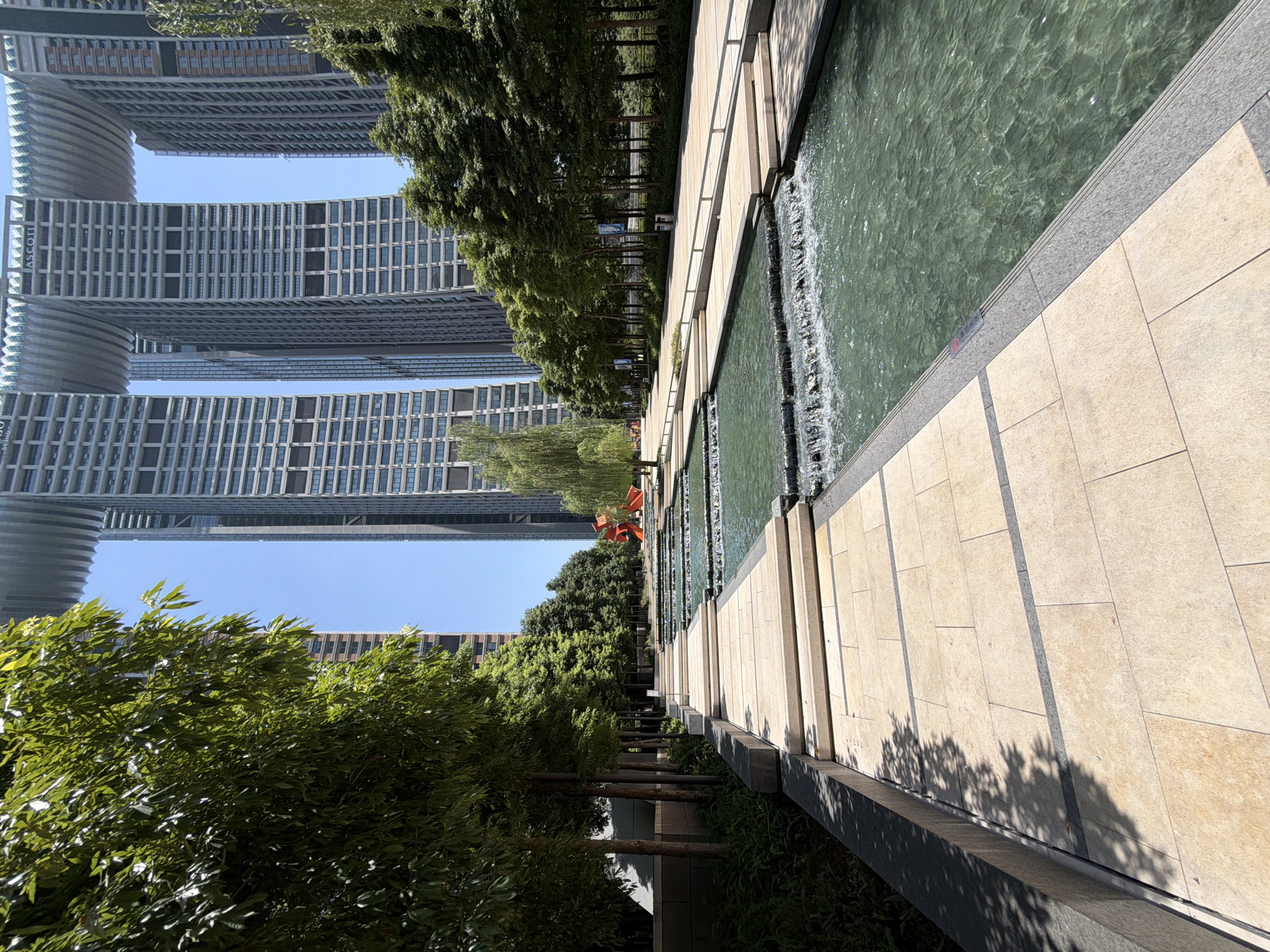 A wide-angle, eye-level shot captures a modern urban landscape featuring a cascading water feature and towering skyscrapers. The foreground is dominated by a light-colored, tiled walkway with darker gray borders, leading the eye towards a series of tiered pools with flowing water. Lush green trees line the left side of the walkway, casting dappled shadows on the tiles. In the mid-ground, a bright red abstract sculpture stands out against the greenery. Beyond the water feature and trees, several contemporary skyscrapers rise into a clear blue sky. These buildings feature distinctive architectural elements, including cantilevered sections resembling bridges connecting the upper floors. The overall scene suggests a well-maintained public space designed for aesthetics and recreation within a bustling city. The location is the Raffles City Chongqing in Yuzhong District, China.