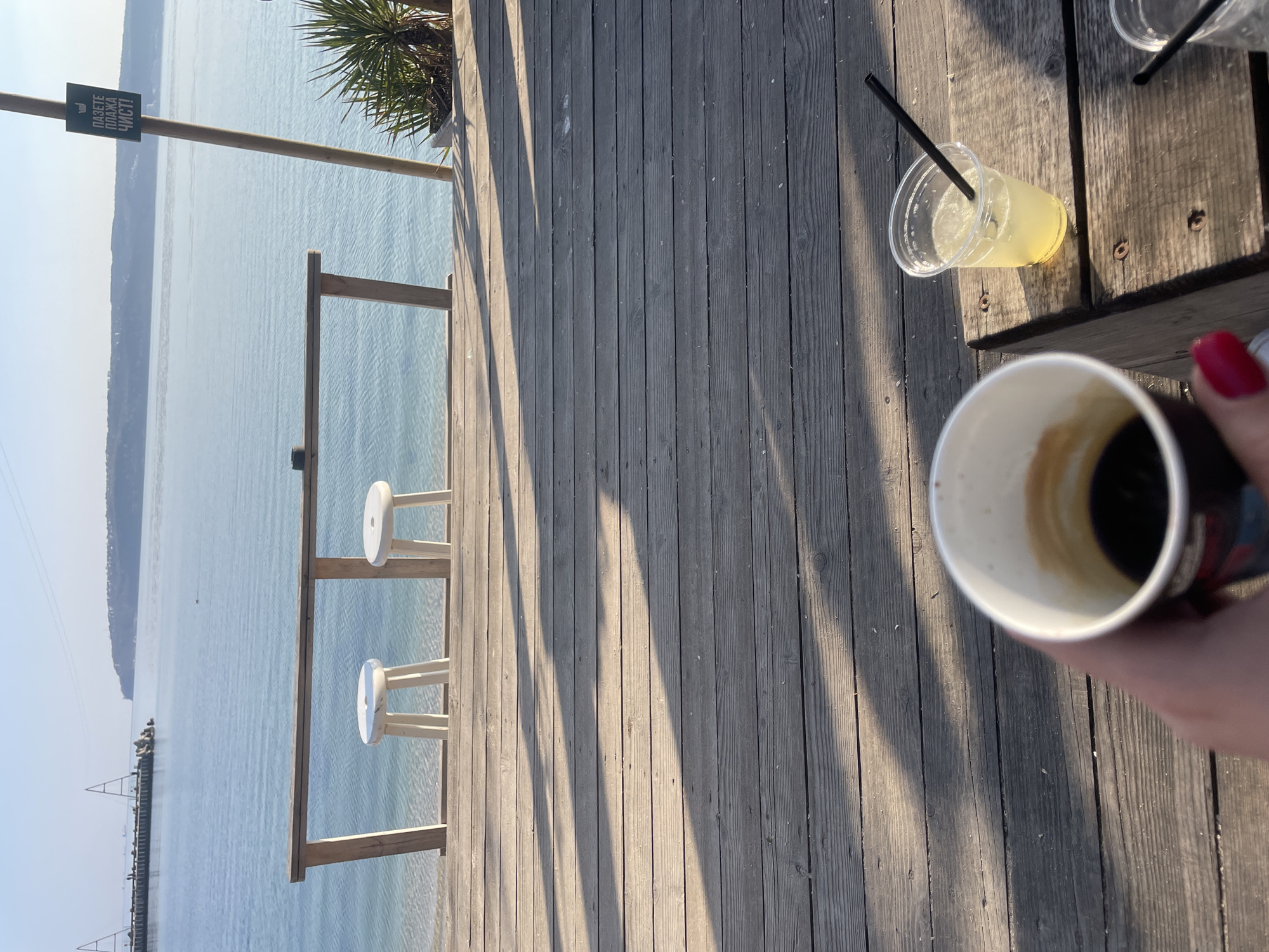 The scene is a wooden deck overlooking the sea in Varna, Bulgaria. On the deck are two white stools facing the sea, a sign in Bulgarian, a palm tree, a table with a lemonade drink and a coffee cup with red nail polish in view. The water has a pier with many boats in the distance. The environment is sunny and bright.