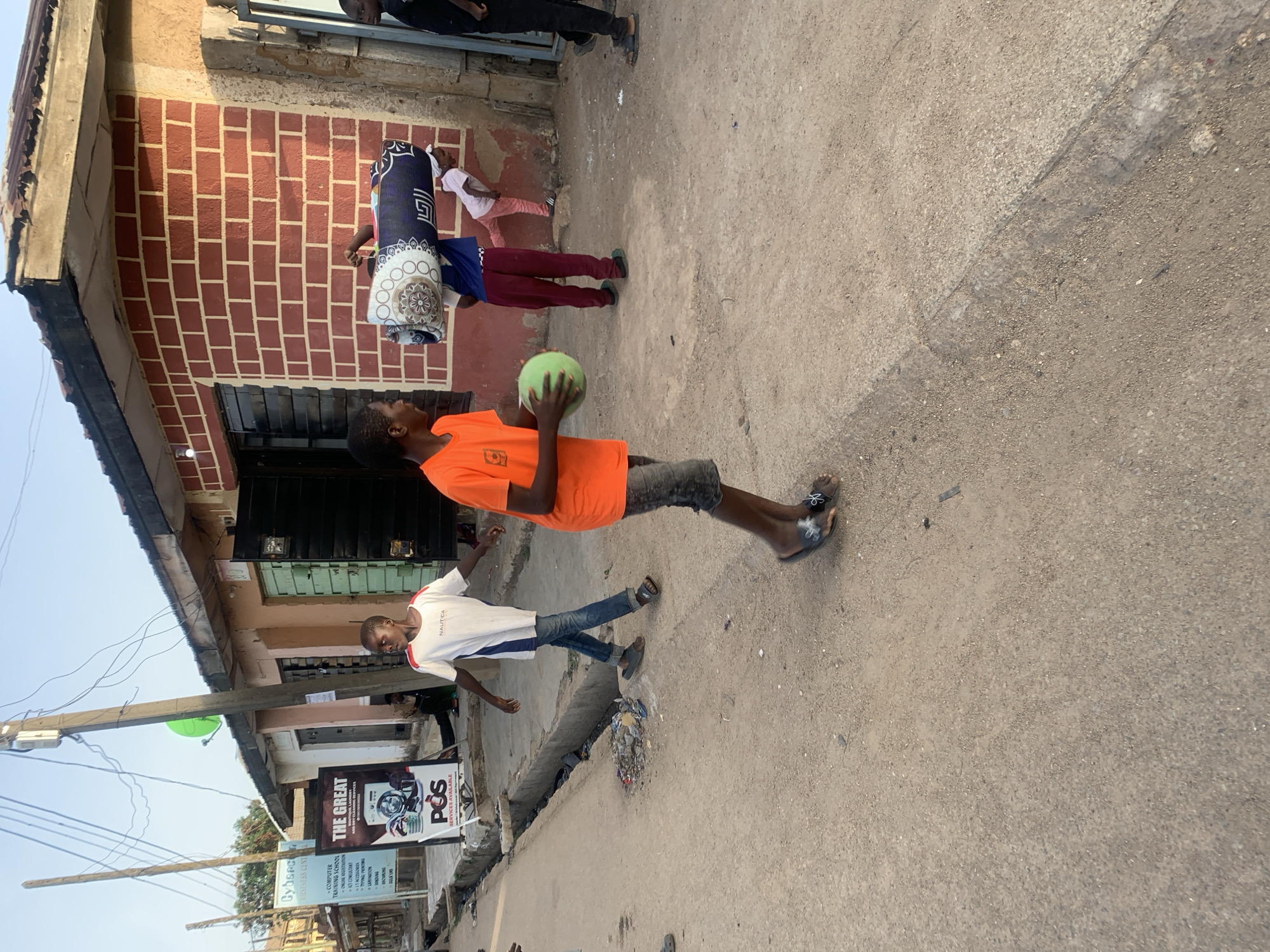 A street scene in Jos, Nigeria. Four children are present. One child in an orange shirt and dark shorts stands with a green ball in his hands. Another child wearing a white t-shirt and jeans is nearby. Two other children are further away, one wearing a blue top and red pants carries a rolled-up item on their back. The other child is wearing pink. Buildings line the street, and there are signs posted, including one for a "POS" (Point of Sale) service. A street light pole is on the left side of the image.