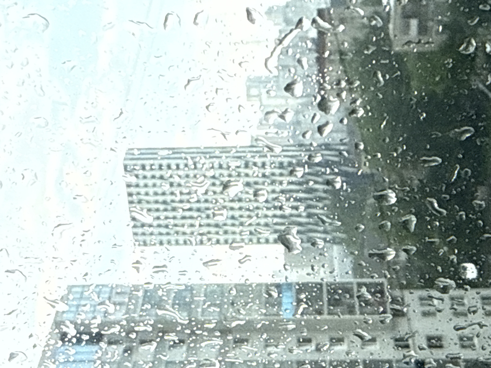 The image shows a cityscape seen through a rain-covered window. Several buildings are visible in the background, with one featuring a distinctive pattern of regularly spaced elements, likely windows or balconies. The foreground is dominated by raindrops clinging to the glass, distorting and obscuring the view. The overall impression is of a rainy day in an urban environment. Identifying the specific location in Yekaterinburg is not possible without further information or visual landmarks within the scene.