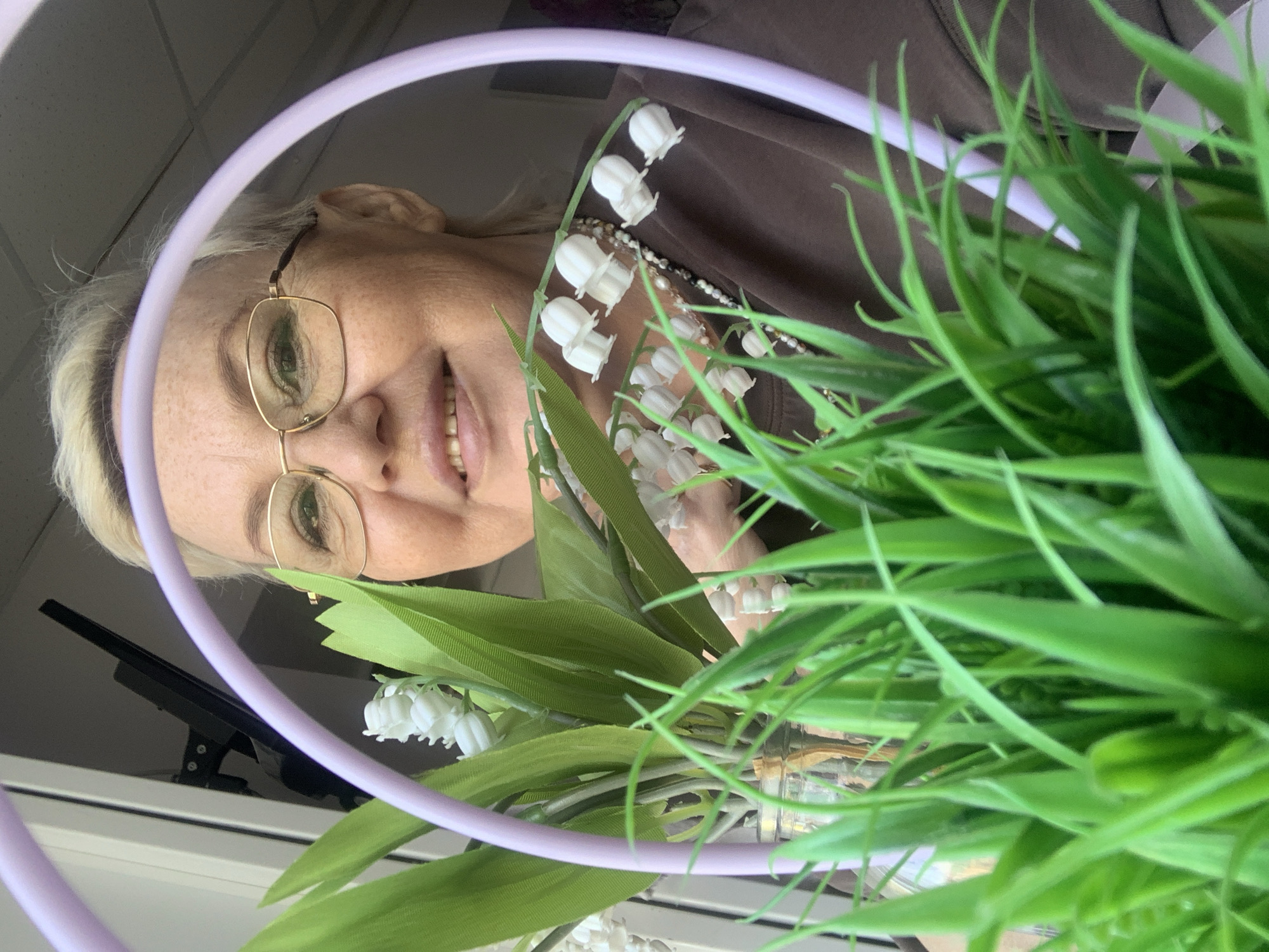 A woman wearing glasses and a brown shirt is holding a bouquet of lily of the valley flowers with green grass. She is positioned in front of a light lavender ring light and appears to be smiling. A window with a dark object that could be a monitor or television screen is visible behind her. The exact location in Chaikovsky, Russia, cannot be determined from the image alone.