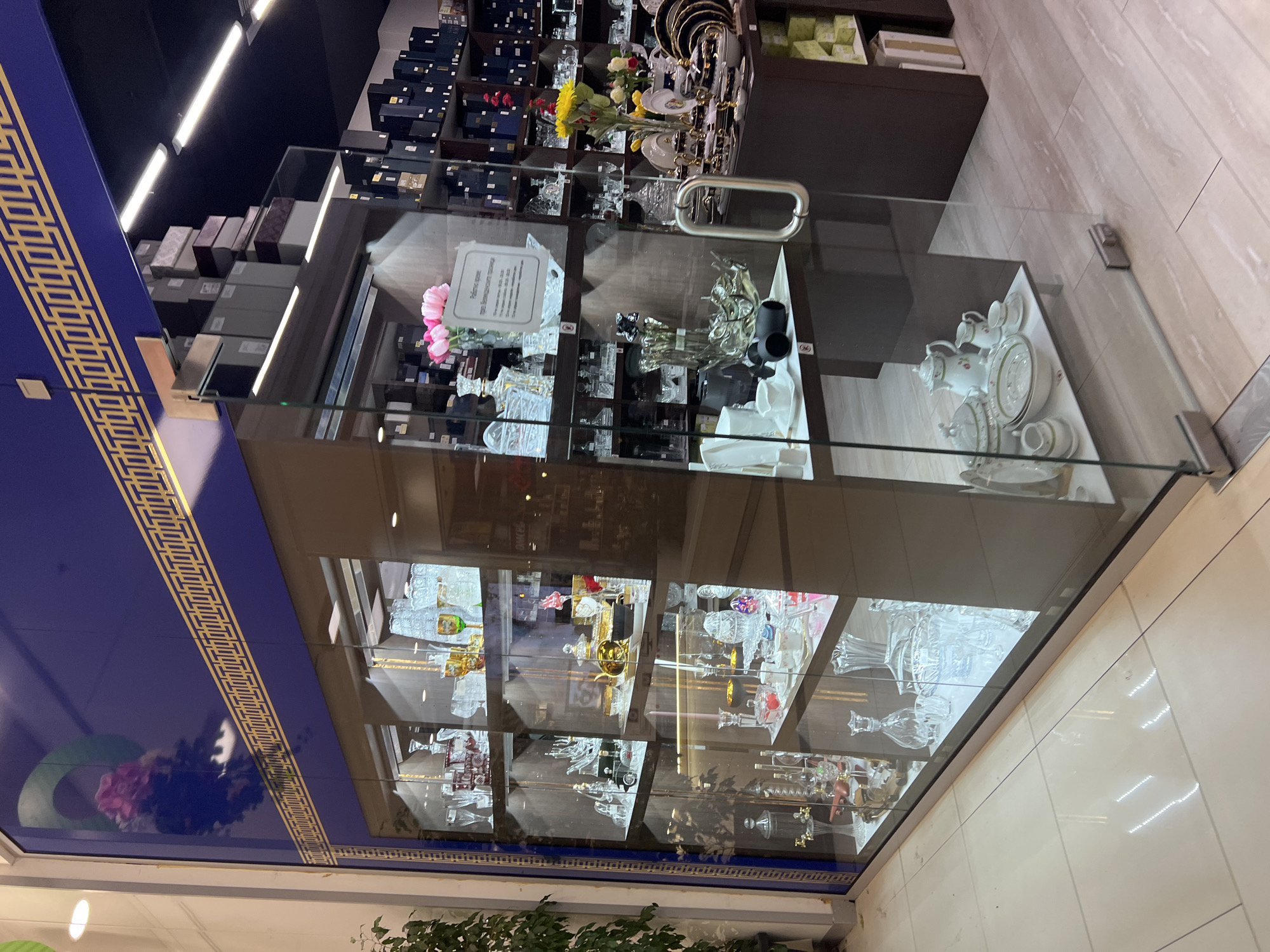 A glass display case filled with various crystal and porcelain items is located in a shop in Plovdiv, Bulgaria. The case has multiple shelves displaying different items like glassware, teacups, plates, decorative objects and flower arrangements. The background shows shelves stocked with boxes and other items for sale. The case is illuminated by interior lighting, emphasizing the glassware. The shop has a dark blue ceiling, with decorative trim and floor tiles that have a light-colored pattern.