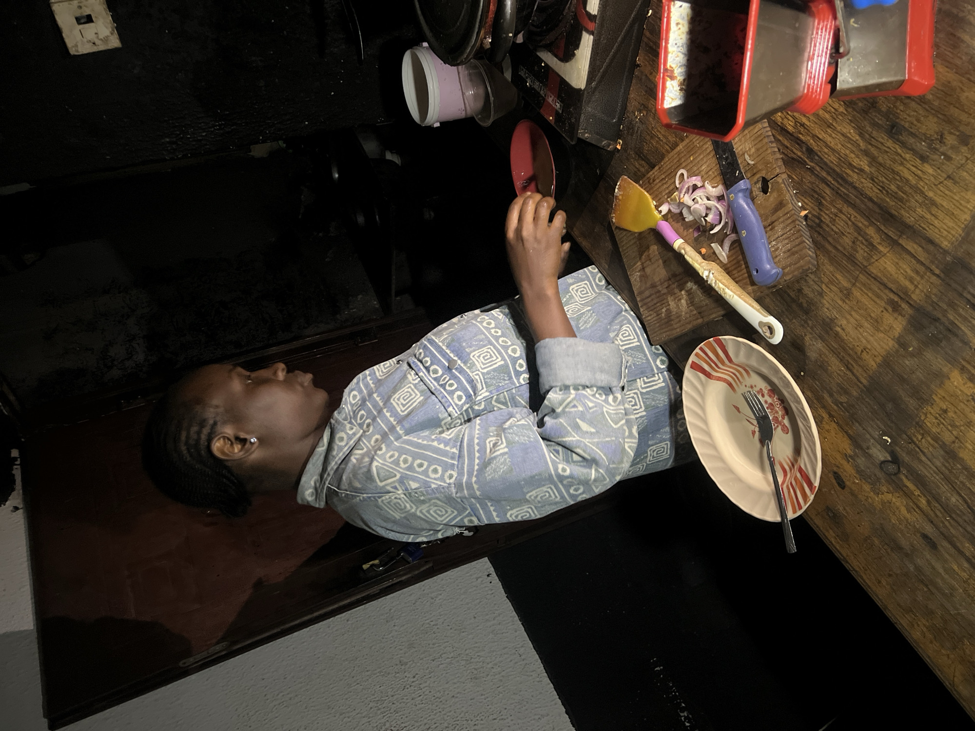 In Durumi, Nigeria, a young woman with braided hair is captured in a dimly lit kitchen. She is wearing a blue denim jacket with a white geometric pattern. Her right hand is outstretched, holding a red plate over a gas stove. On the wooden countertop in the foreground, a white oval plate with orange and red stripes lies beside a fork. A yellow spatula, sliced red onions, and a blue-handled knife are arranged on a cutting board. To the right, a red and black food processor sits on the counter. The background is dark and appears to be the kitchen wall and shelving, with a pink bucket visible on a shelf. The overall scene suggests food preparation is in progress.