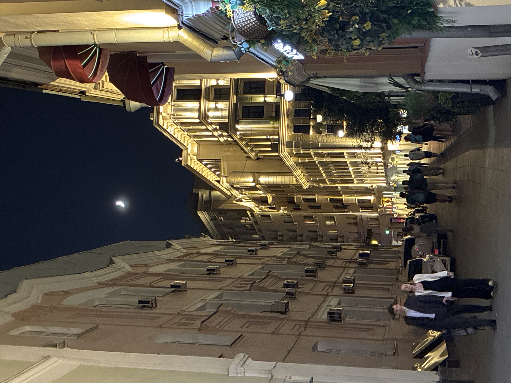 The image depicts a Moscow street at night. Pedestrians stroll along the sidewalk. Buildings are illuminated with many lights. A crescent moon is visible in the dark sky.