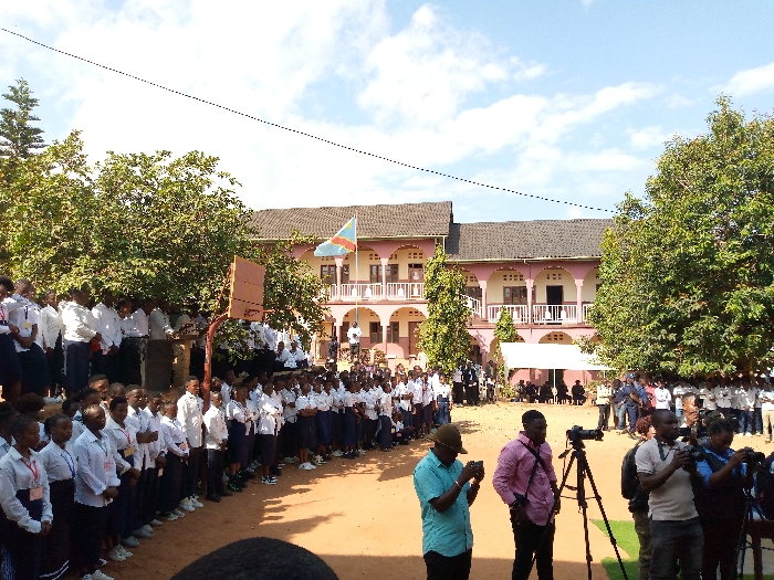 A large group of people, predominantly young adults, stand in a schoolyard in Beni, Democratic Republic of Congo. The group is organized in rows, facing a white tent with a small group of people seated inside. Several individuals wearing white shirts and black trousers are standing in front of the group of adults. Two trees, one on each side of the frame, provide shade for the people gathered in the yard. There are several cameras set up on tripods with photographers taking pictures. A basketball hoop is attached to a pole near the front of the group of adults, in front of the building. The building in the background has a large, curved portico and a single Congo flag flying above the building. The sky is clear and blue with a few scattered white clouds. The ground is brown and dusty. The scene is a formal gathering, likely a graduation ceremony, or a similar event. The environment is hot and sunny with a dry, dusty atmosphere. The location is in the city of Beni, Democratic Republic of Congo, at the coordinates (latitude: 0.492, longitude: 29.483).