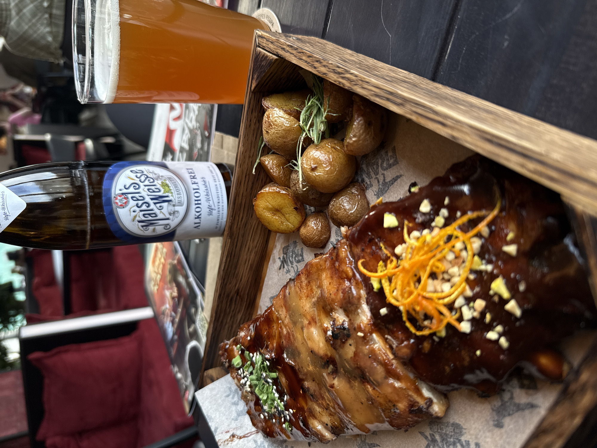 This close-up, top-down image captures a meal served in what appears to be a restaurant or bar setting in Krasnoyarsk, Russia. The central focus is a generous portion of BBQ ribs, glistening with a dark, savory glaze. The ribs are artfully garnished with finely chopped green onions or chives and white sesame seeds on one end, and on the other, bright orange zest strips along with small, light-colored nuts or seeds scattered across the sauce.
Accompanying the ribs are several small, whole roasted potatoes, their skins golden brown and slightly crispy. These potatoes are adorned with a sprig of fresh rosemary. Both the ribs and potatoes are presented inside a rustic wooden tray or box, lined with brown paper that has faint, illegible text, possibly restaurant branding like "FRANK."
To the left of the food, an amber-colored glass of beer with a frothy head is visible, indicating a refreshing beverage. Below the glass, a brown glass bottle of "Maisel's Weisse" beer is lying horizontally. The label clearly states "ALKOHOLFREI" (non-alcoholic) and "Hefe-Weissbier" (yeast wheat beer), along with "AUS BAYREUTH" and "FAMILIENBRAUEREI GEGR. 1887," suggesting a German origin.
The background shows a dark tabletop, possibly made of slate or chalkboard, with some faint marks. Reflections in the background include blurred red cushioned seating and a portion of a poster or painting, adding to the ambiance of a casual dining establishment. The lighting suggests it is daytime, providing clear visibility of the food's appealing textures and colors. No people are visible, and the scene implies a meal ready to be enjoyed.