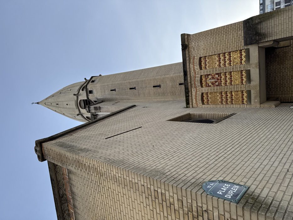 A low-angle shot captures a tan brick building and a taller, cylindrical tower against a bright sky. Inscriptions adorn the building's side, adjacent to an open doorway. A blue street sign labeled "Place Dupleix" is visible near the bottom corner. The architecture features a consistent brick pattern throughout, with minimal ornamentation beyond the inscriptions and tower's form.