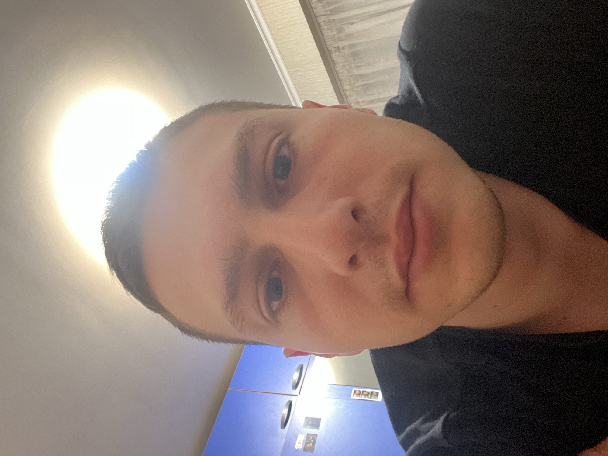A selfie of a light-skinned male with short brown hair, blue eyes, and stubble is presented. He is wearing a black shirt. The photo is taken at an upward angle, with a bright light source above. The background includes a light-colored ceiling and blue cabinets with silver handles. Small items are affixed to the cabinets. The location is unidentifiable as Burgas, Bulgaria, based solely on the visual information provided.