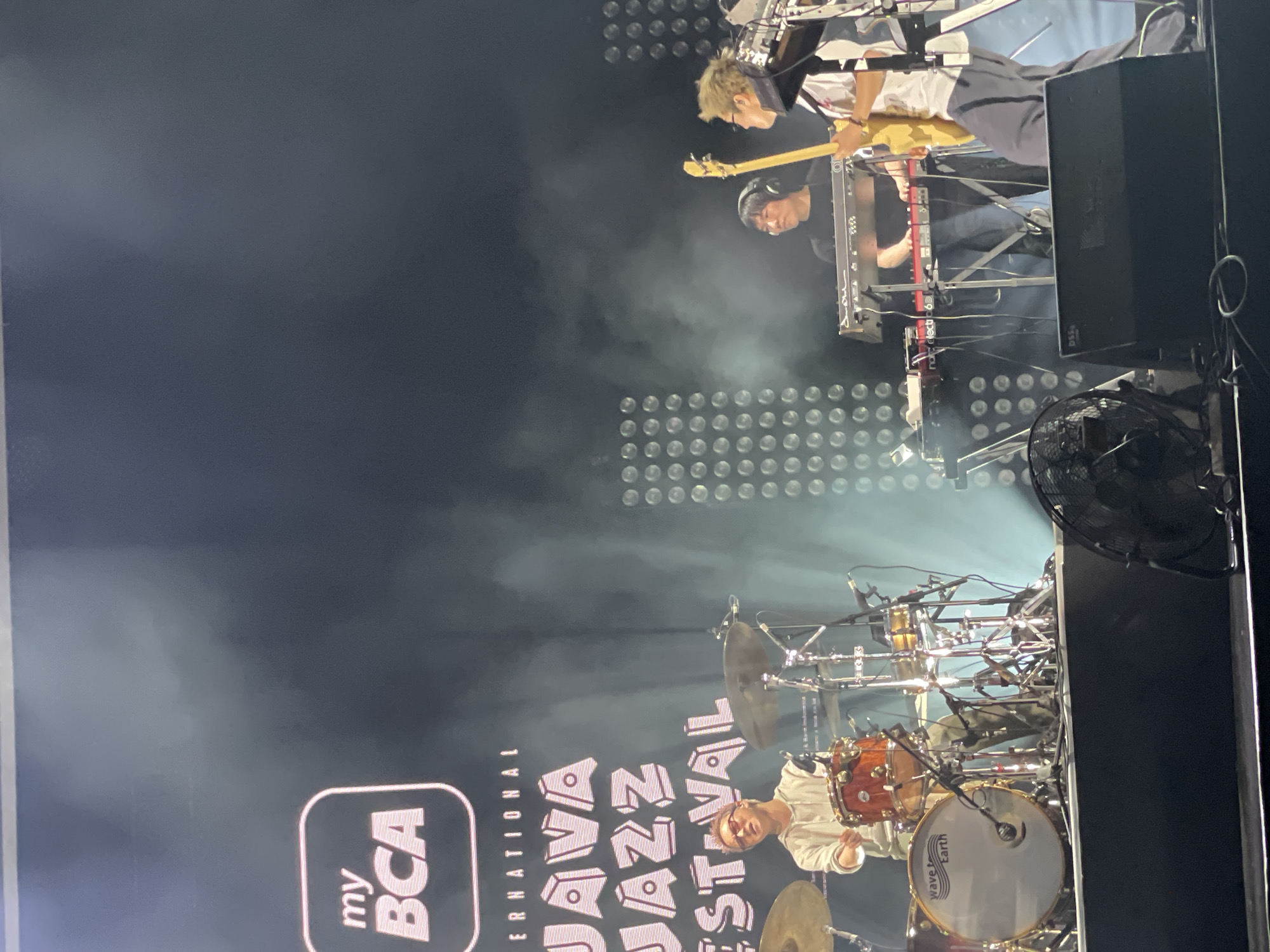The image depicts a live musical performance at the Java Jazz Festival, presumably the BCA International Java Jazz Festival. The scene is likely set in Salembaran, Indonesia, based on the festival's name and origin. Three musicians are visible: a drummer, a keyboardist, and a guitarist/bassist. The drummer is seated behind a drum set, playing. The keyboardist is operating a red keyboard, and the guitarist is playing a light-colored guitar. Behind the performers, there is a wall of lights, and fog is present on stage. The "my BCA" logo is visible on the left side of the image, along with partial views of the "JAVA JAZZ FESTIVAL" banner. A black fan is visible stage right.