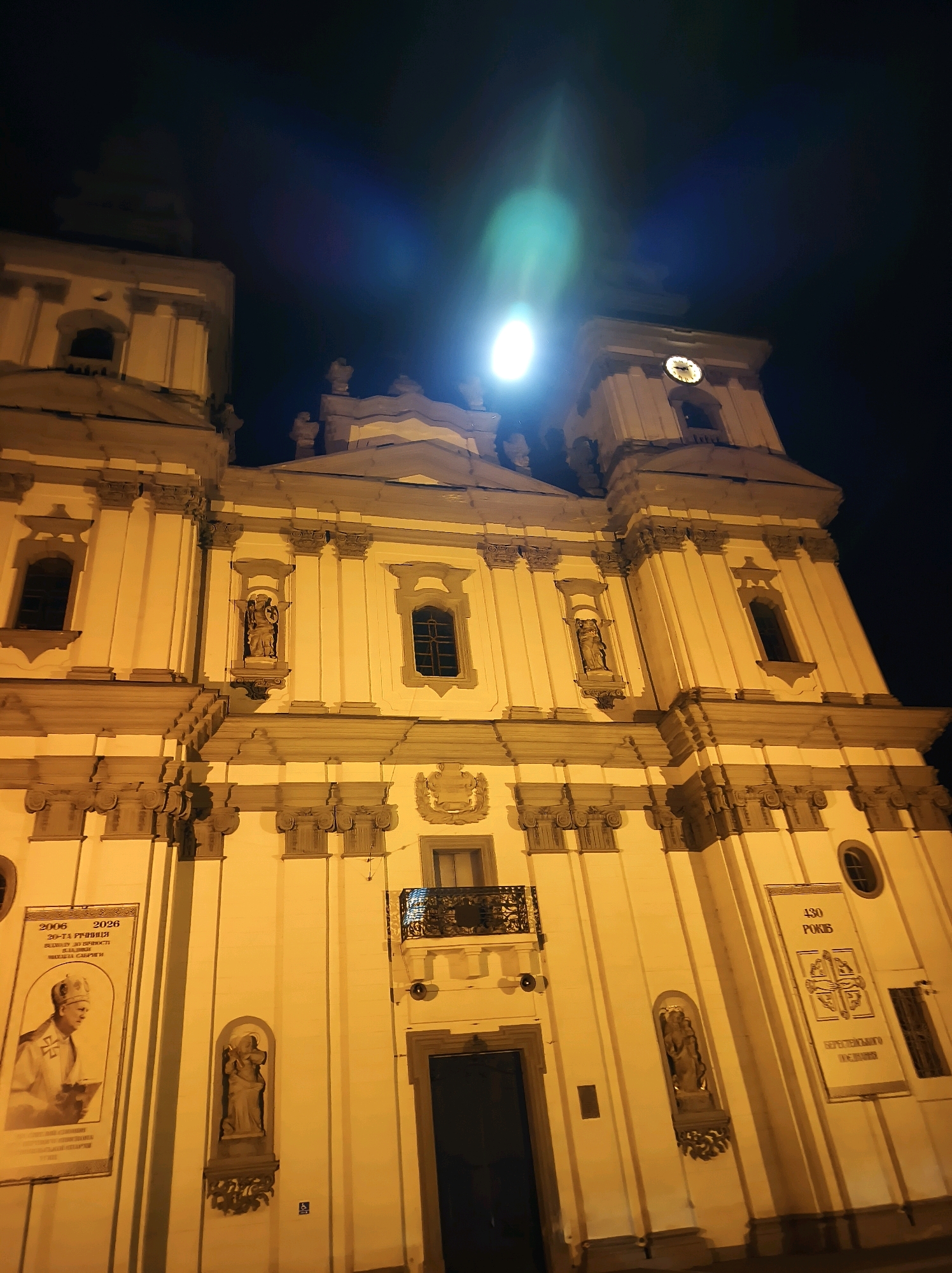 The image is a nighttime, low-angle shot of the Ternopil Cathedral (Nativity of the Blessed Virgin Mary) in Ternopil, Ukraine. The facade of the church is illuminated by yellow artificial lighting, casting long shadows. Two prominent towers flank the central structure, one of which features a clock face. Various architectural details such as columns, windows, and statues are visible. Two large banners are displayed on the facade. The left banner shows a portrait of a man in religious attire and text in Cyrillic script. The right banner displays a symbol and more Cyrillic text. The sky above is dark, with a bright, lens-flared light source visible, likely the moon or a strong artificial light, causing a green and blue light streak. The overall impression is of a grand, historical building at night.