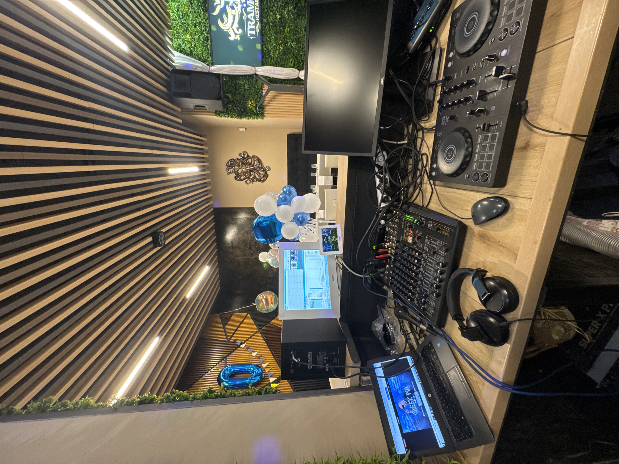 The image depicts a DJ setup in what appears to be a restaurant or event venue. The setup includes a laptop, a DJ controller, a mixer, a monitor, and headphones, all arranged on a wooden counter. In the background, there is a wall with decorative wooden panels and a large green wall with artificial plants. A "Tramonti Restaurant" sign is visible on the green wall. Balloons, including a large blue "0" balloon, are also present, suggesting a celebration or party. The ceiling is adorned with wooden slats and strip lighting. The overall atmosphere is festive and professional, indicating a space prepared for entertainment. The exact location in Isperih, Bulgaria cannot be determined from the image alone.