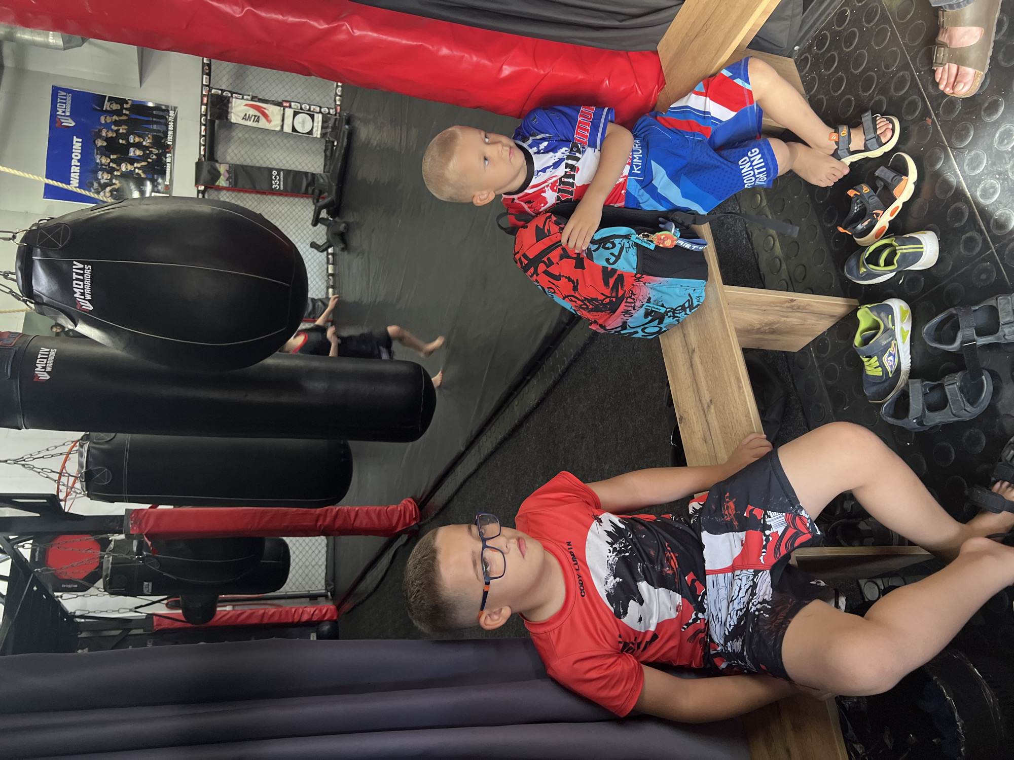 This image captures an interior scene of a martial arts or combat sports gym, likely in Sochi, Russia, based on the provided context and visible Russian text. Two young boys are prominently featured, resting on wooden benches or risers.
The boy in the foreground, wearing glasses, a red and black short-sleeved shirt with the text "CODY LUND IN", and matching shorts, is lying down on a padded surface, looking towards the left. Behind him, another boy with blonde hair, dressed in a blue, white, and red athletic shirt and shorts (featuring "KIMURA", "ROUND", and "FIGHTING" text), sits upright with a colorful backpack next to him. Several pairs of shoes and sandals are scattered on the dark, textured floor beside them, and an adult's feet are also visible.
The setting is characterized by typical gym equipment: large black punching bags, some branded "MOTIV WARRIORS", hang from the ceiling. A fighting cage structure, with red padded rails and black mesh fencing, is visible in the background, including banners with "ANTA" and "33°" logos. A large blue poster on the wall displays "MOTIV WARRIORS" and "WARPOINT", along with Russian text "Вишня 10/5819291 854-77-00" (likely an address and phone number for the gym). In the deeper background, other individuals are partially visible, suggesting training or activity in the cage area. The overall atmosphere appears to be a break or observation period during a training session.