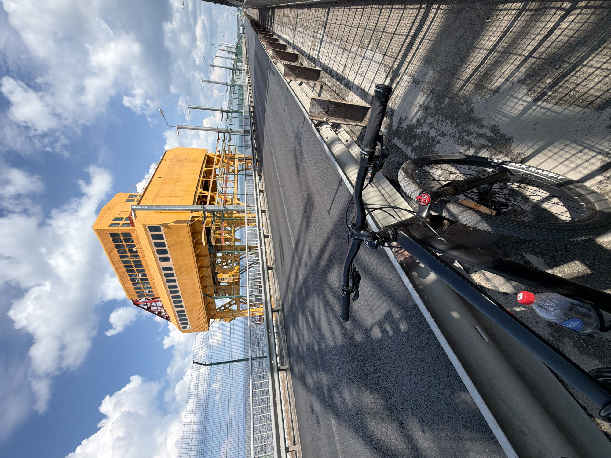 The photograph is taken from the perspective of a cyclist on a bridge, looking towards a large, bright yellow industrial structure. The structure appears to be a control room or observation post, with numerous windows lining its upper section. It is supported by a framework of yellow and red metal beams. A chain-link fence runs along the edge of the bridge, separating the road from the water beyond, which is calm and reflects the sky. The bridge itself is paved with asphalt and has a pedestrian walkway on the right side, characterized by a concrete surface and a metal railing. Streetlights are visible along the bridge. The sky is blue with scattered white clouds, and the sun is casting long shadows across the bridge. In the foreground, the front wheel, handlebars, and part of the frame of a black mountain bike are visible. A partially filled plastic water bottle with a red cap rests in a bottle cage on the bike's frame. The scene is set on the Chaikovsky Bridge, located in the city of Chaikovsky, Russia, spanning the Kama River.