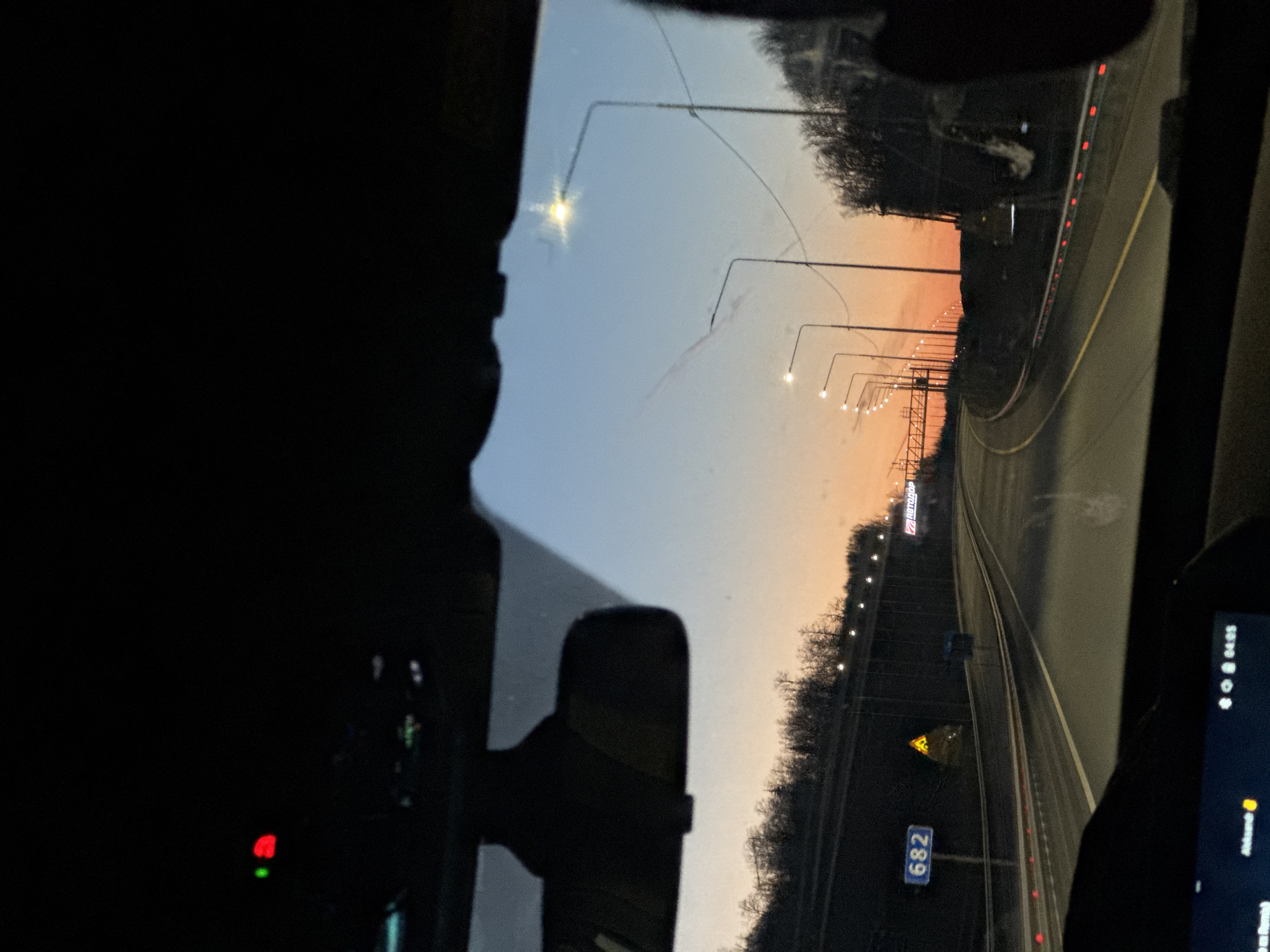 A nighttime view from inside a vehicle moving along a highway. Streetlights illuminate the road and roadside trees. A road sign with the number "682" is visible on the right side of the road. The sky transitions from a dark blue to orange towards the horizon, likely indicating either sunrise or sunset. A dashboard screen is partially visible, showing time information. The city and state are unknown.