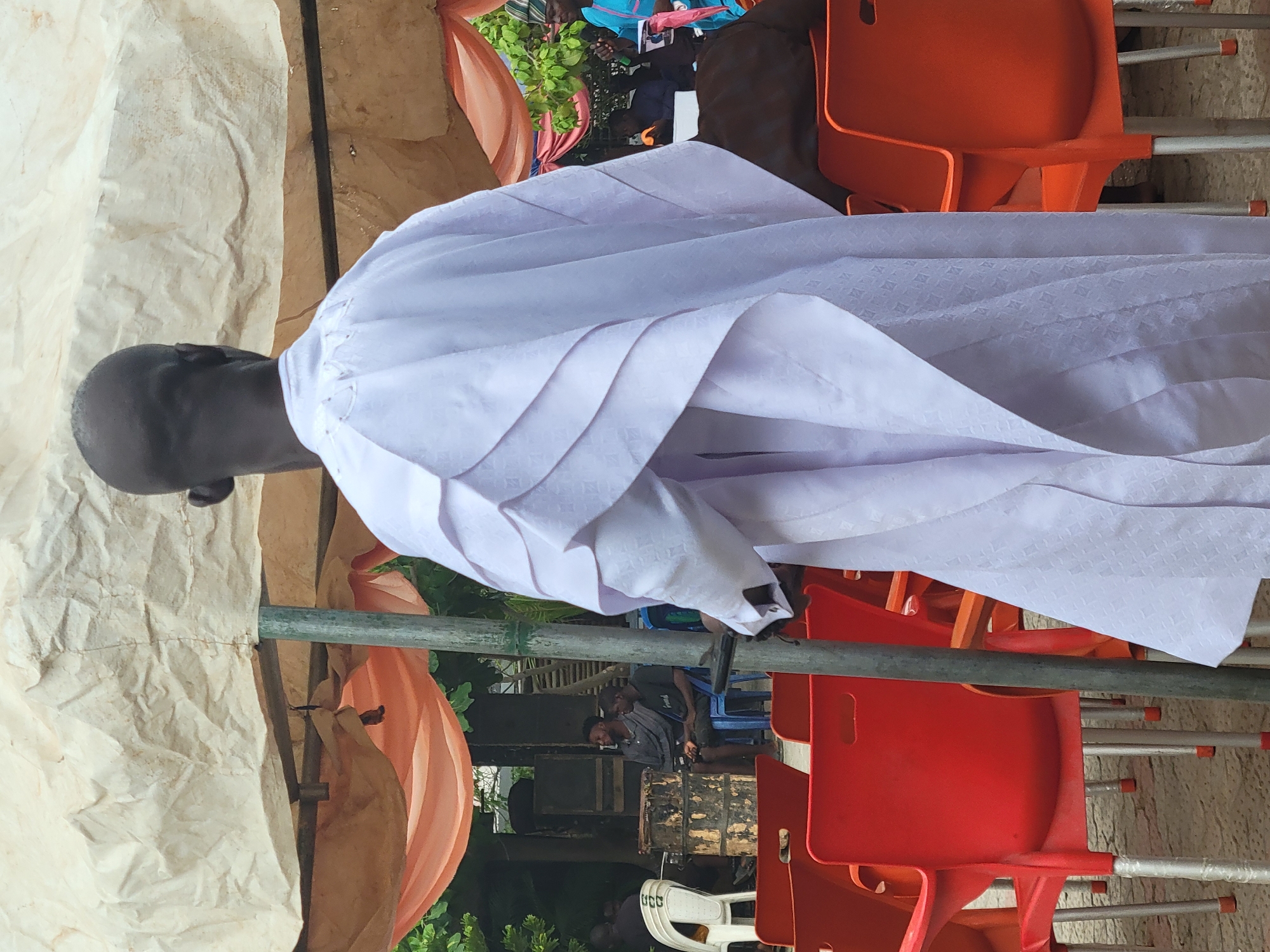 An exterior shot depicts a man in a white robe standing beside a stack of orange chairs. The setting appears to be under some sort of draped tent or canopy. The location is Suleja, Nigeria.