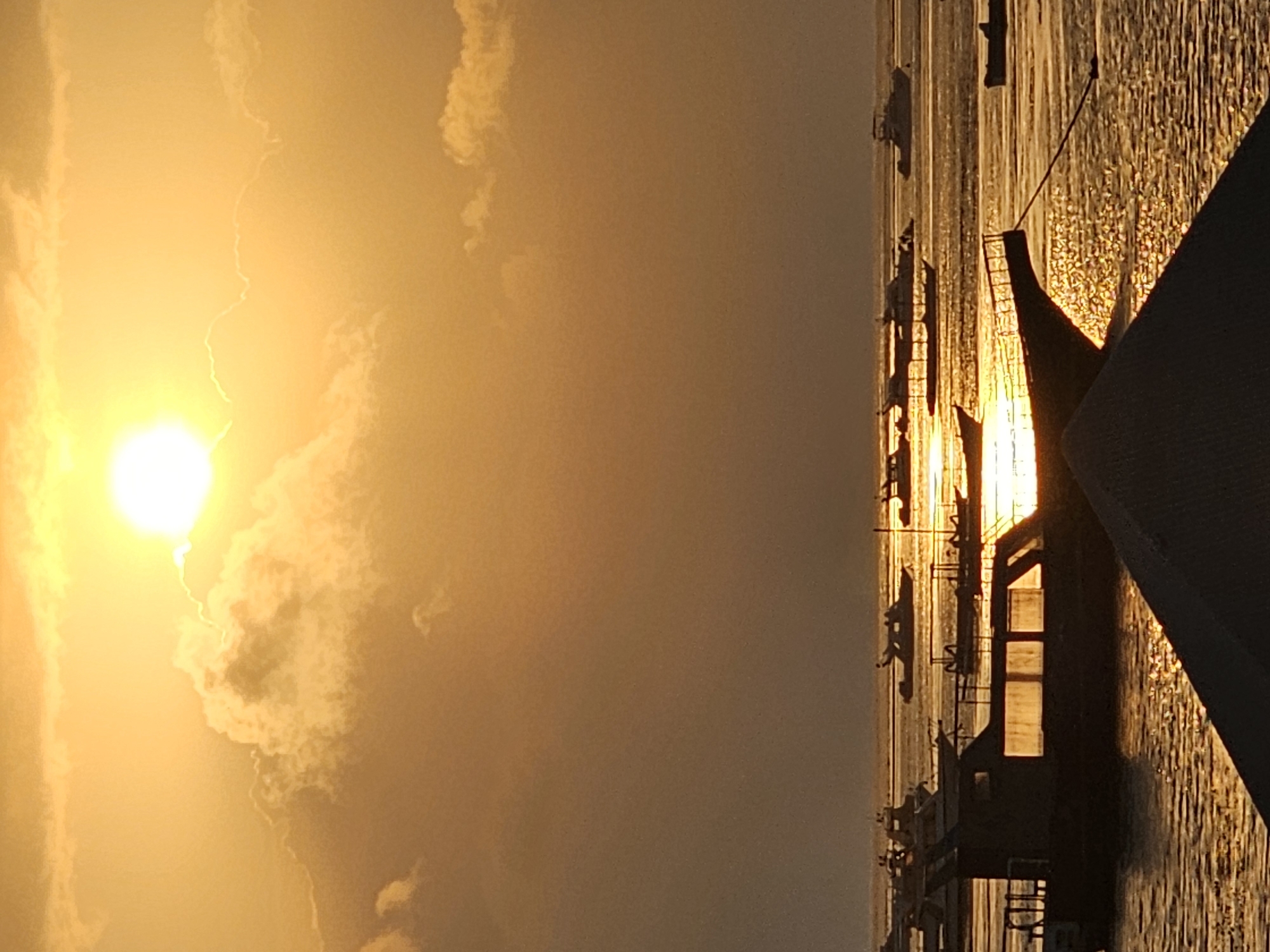 The image depicts a sunset over the ocean in Denpasar, Indonesia. The sky is a vibrant orange and yellow, with the sun partially obscured by clouds. Several boats are scattered across the water, some of them silhouetted against the setting sun. The water itself is calm and reflects the warm colors of the sky. In the foreground, a portion of a dark, angled roof is visible, suggesting a viewpoint from a building or elevated structure. The overall scene is serene and picturesque, characteristic of a tropical sunset.