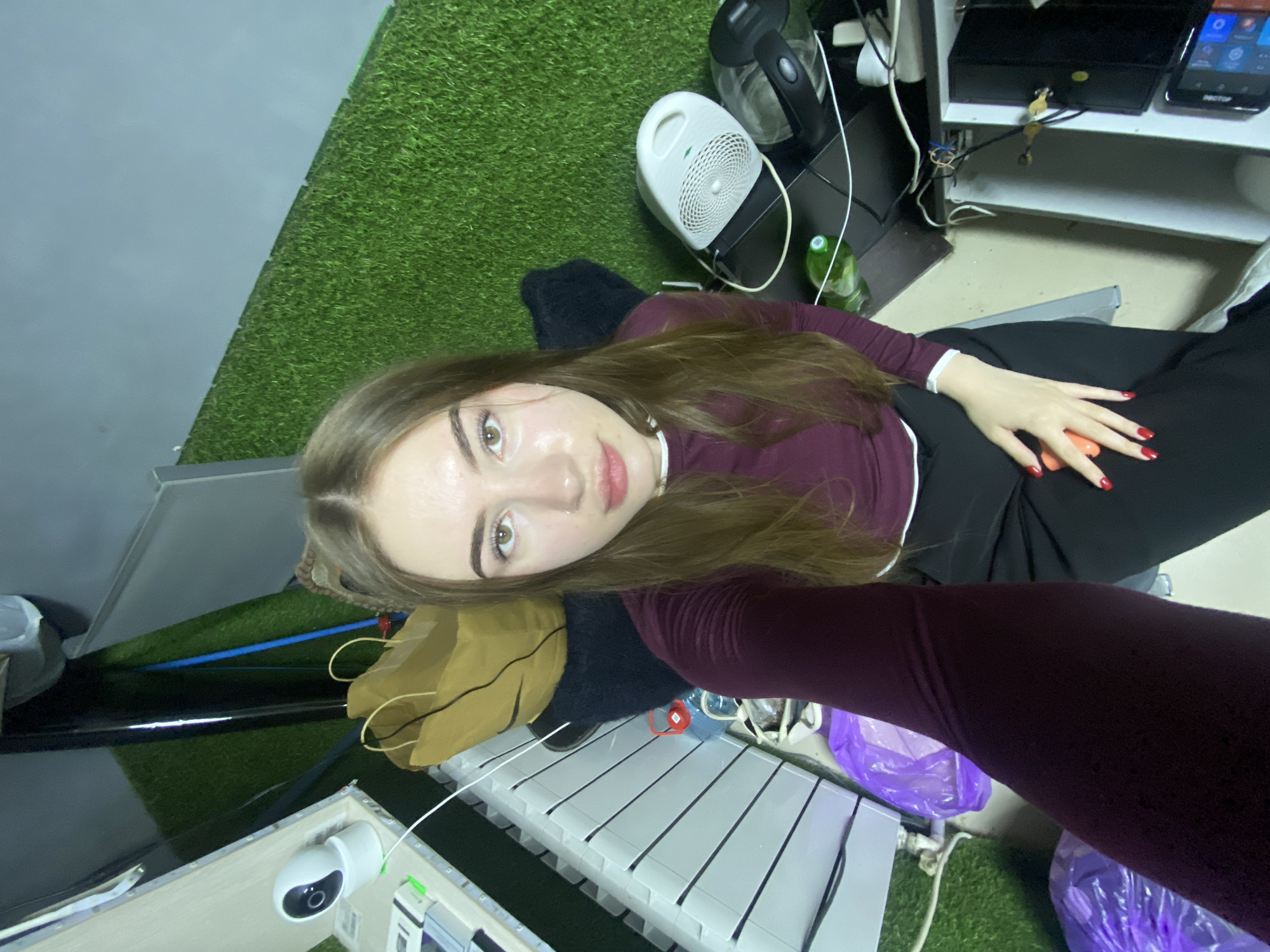 A young woman with long brown hair, fair skin, and light eyes looks directly at the camera from a high-angle perspective. She wears a dark maroon long-sleeved top with a white collar or trim and black pants. Her fingernails are painted red. Her left hand rests on her lap.
The setting is an indoor space with artificial green turf covering the floor, creating a distinctive ground surface. A plain gray wall is visible in the upper left of the image. The environment appears functional and somewhat cluttered, featuring a mix of electronic devices and household items.
To the woman's right, on a black shelf or table, there is a white portable fan or heater, a clear electric kettle, and a green plastic water bottle. Various cables and wires are visible, alongside a black electronic box and a smartphone or small tablet displaying colorful app icons. Keys are seen hanging from a hook on the shelf.
To the woman's left, a white security camera is mounted on a light-colored surface. Below it, a white slatted panel, possibly a radiator, is visible. Other items in this area include a black surface, a brown paper bag, a blue pipe or cable, and purple plastic bags on the artificial turf. A black furry object, possibly a cushion or pet bed, is situated near her.
The overall impression is that of a casual, multi-purpose, or possibly utility-oriented room, characterized by the unusual artificial grass flooring and the blend of personal and electronic equipment. The time of day is not discernible due to indoor lighting. No clear legible text is visible in the image.
