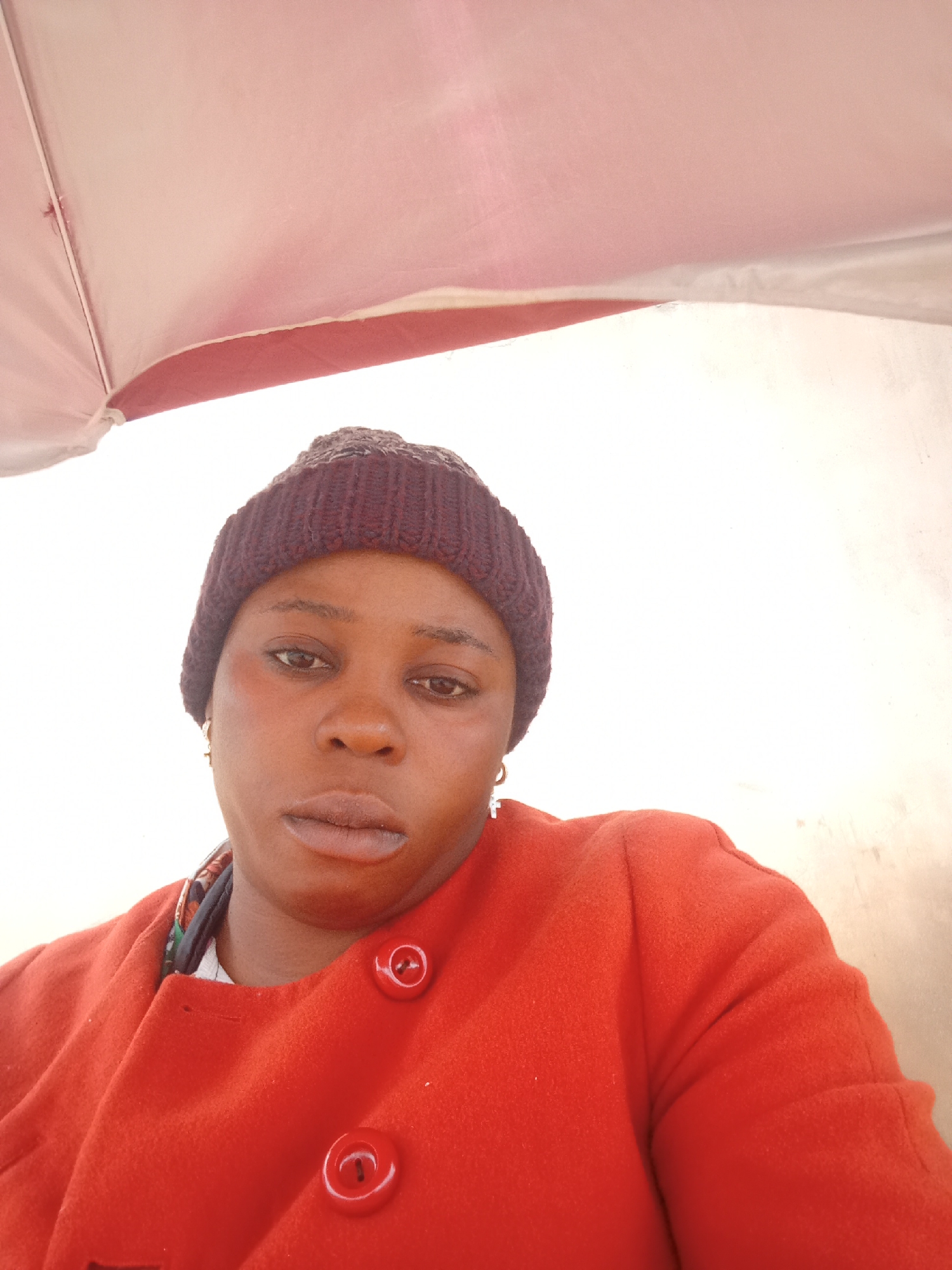 A medium shot captured from a slightly low angle shows a Black woman from the chest up, wearing a dark knit beanie and a bright orange coat with large, round buttons. Her head is tilted slightly to her left, and she is looking directly at the camera. The background appears to be an outdoor setting, possibly under a pink and white umbrella, with a light-colored wall to the right. The image is well-lit, highlighting the woman's facial features and the texture of her clothing.
