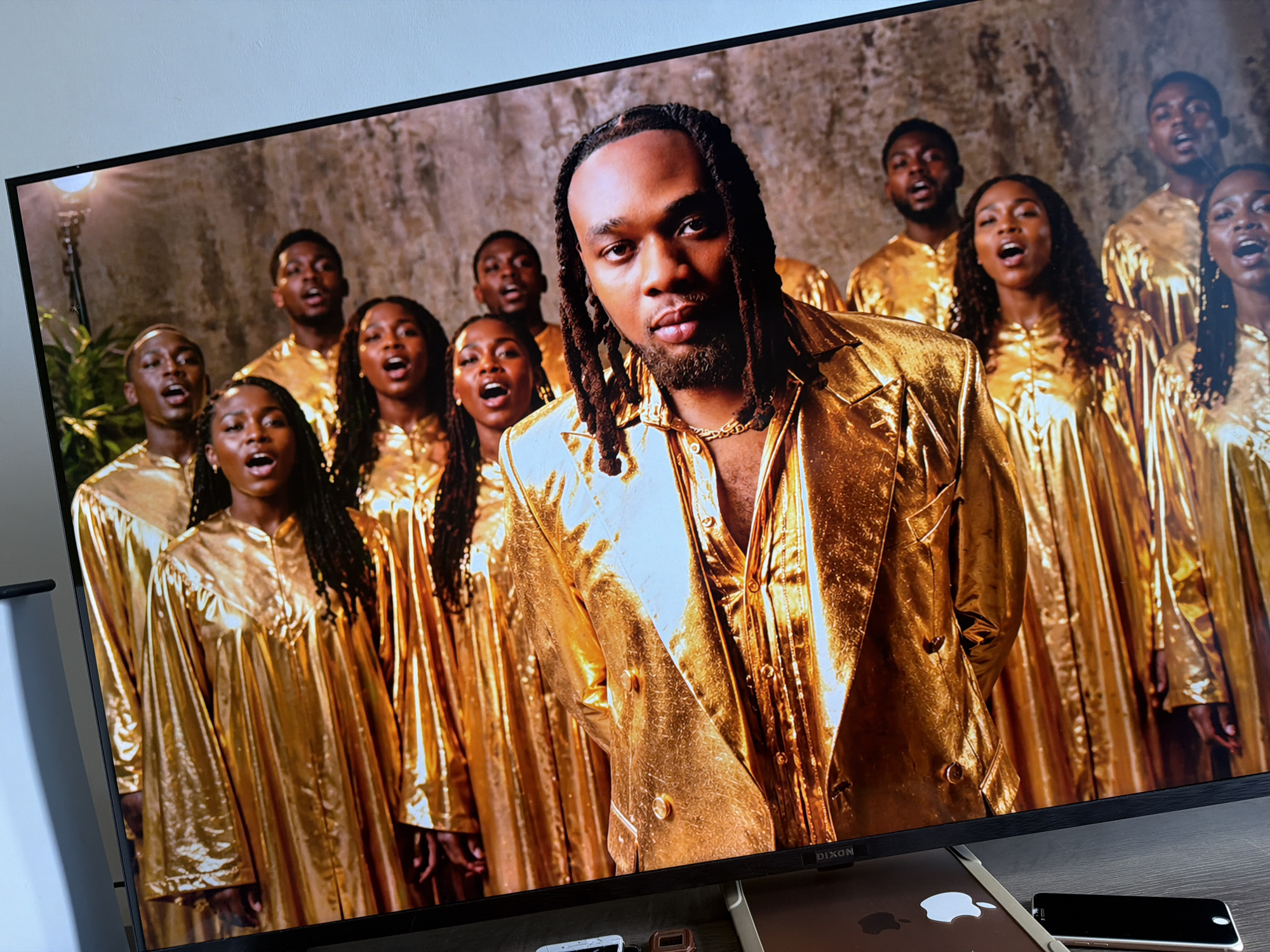 The image captures a vibrant scene displayed on a screen, featuring a group of Black individuals, likely a choir, in striking gold attire. In the foreground, a man with long dreadlocks and a beard stands prominently, wearing a shiny gold suit jacket and matching shirt, accessorized with a gold chain. He looks directly at the viewer with a serious, confident expression.
Behind him, several other men and women are arranged as a choir, uniformly dressed in reflective gold robes or gowns. Most of the choir members have their mouths open in song, conveying a sense of active performance and earnestness. The background consists of a textured, light brown or grey wall, possibly stone or concrete, with a vintage-style lamp fixture visible on the far left. The lighting is warm and dramatic, highlighting the metallic sheen of the gold garments.
The scene suggests a musical performance, possibly a gospel choir or a music video shoot, characterized by its opulent aesthetic. Below the screen, parts of a silver MacBook with an Apple logo and a black smartphone are visible on a dark surface. The brand name "DIXON" is visible on the bottom bezel of the screen. The event takes place in an indoor or controlled studio setting, consistent with the dynamic cultural and artistic environment often found in Cape Town, South Africa.