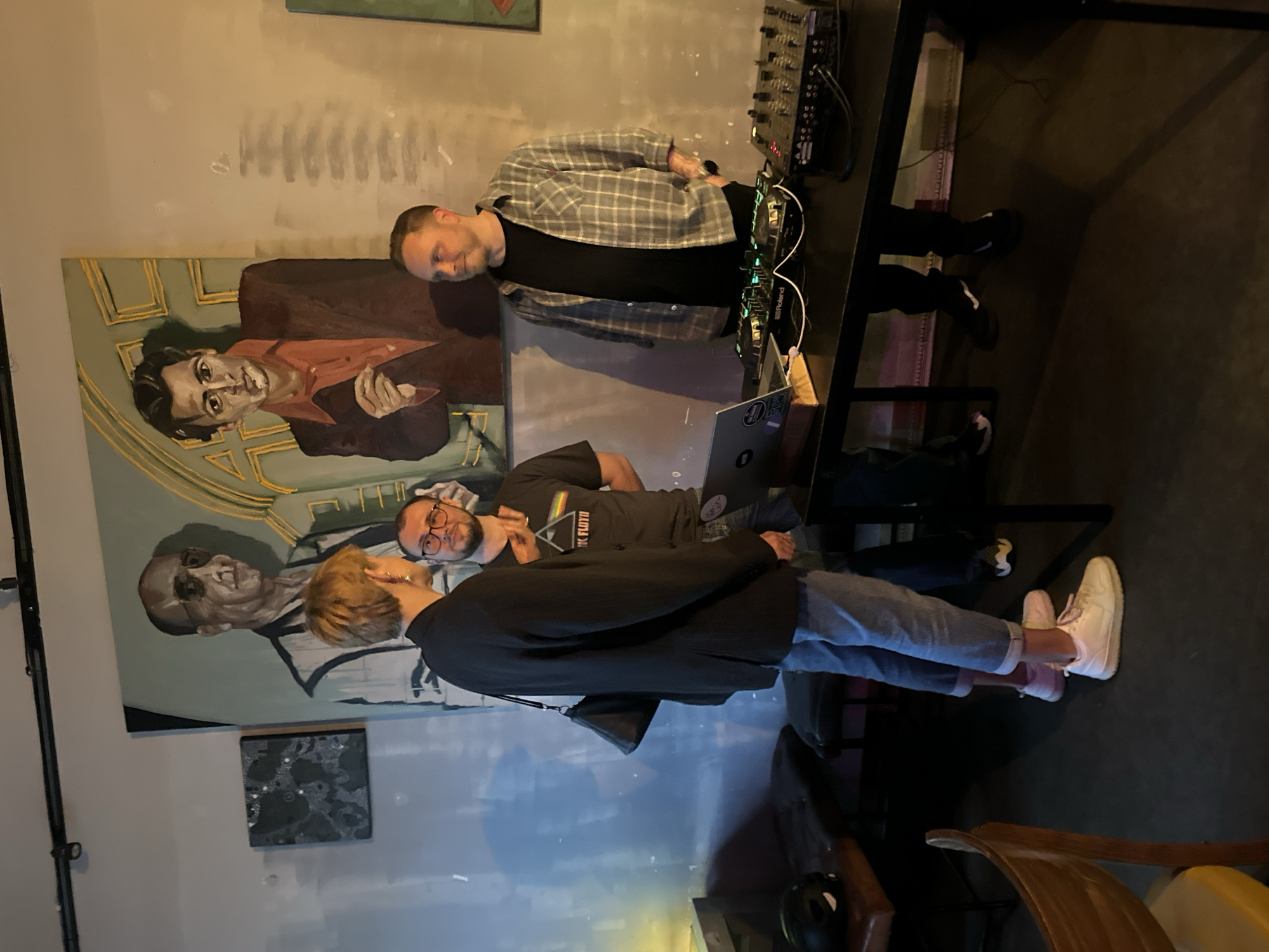 In an indoor setting, likely a bar or club in Riga, Latvia, four individuals are present. Dominating the background is a large, dual-portrait painting of two men, one wearing glasses and a suit, the other in a suit and orange shirt. To the left of the painting hangs a smaller, dark square artwork.
In the foreground, a woman with short blonde hair, wearing a black blazer, blue jeans, and white sneakers, stands facing two men. She carries a black handbag over her shoulder.
One man, wearing glasses and a dark t-shirt with a Pink Floyd logo, stands to her right. He is gesturing with his hands as if in conversation.
To the right of these three, a man in a grey plaid shirt over a black t-shirt and black pants stands behind a black table. On the table are DJ equipment, including two decks and a mixer, as well as a laptop. This man appears to be operating the DJ equipment.
The environment suggests a social gathering, possibly an event or a casual get-together in a dimly lit establishment, with musical equipment implying entertainment. The floor is dark.