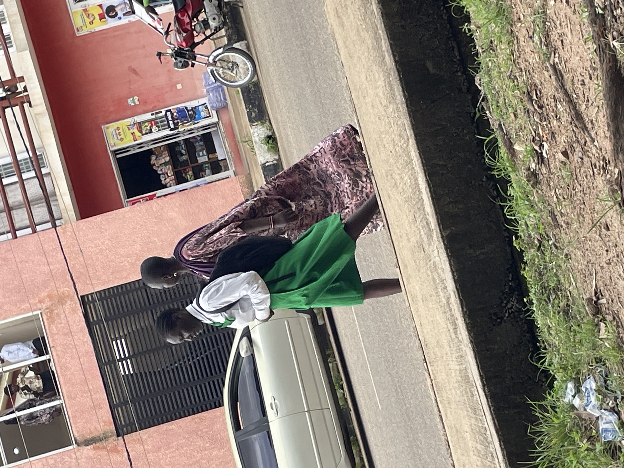 In Jos, Nigeria, two individuals walk on a sidewalk adjacent to a street. The person in the foreground wears a white shirt and a green skirt and carries a black backpack. To their right, another person in a long, patterned purple dress walks alongside. A light-colored car is parked on the street. In the background, a building with red walls features windows and a shop entrance with merchandise displayed inside. A red motorcycle is parked near the shop. The scene appears to be in a commercial or residential area, typical of a Nigerian city.