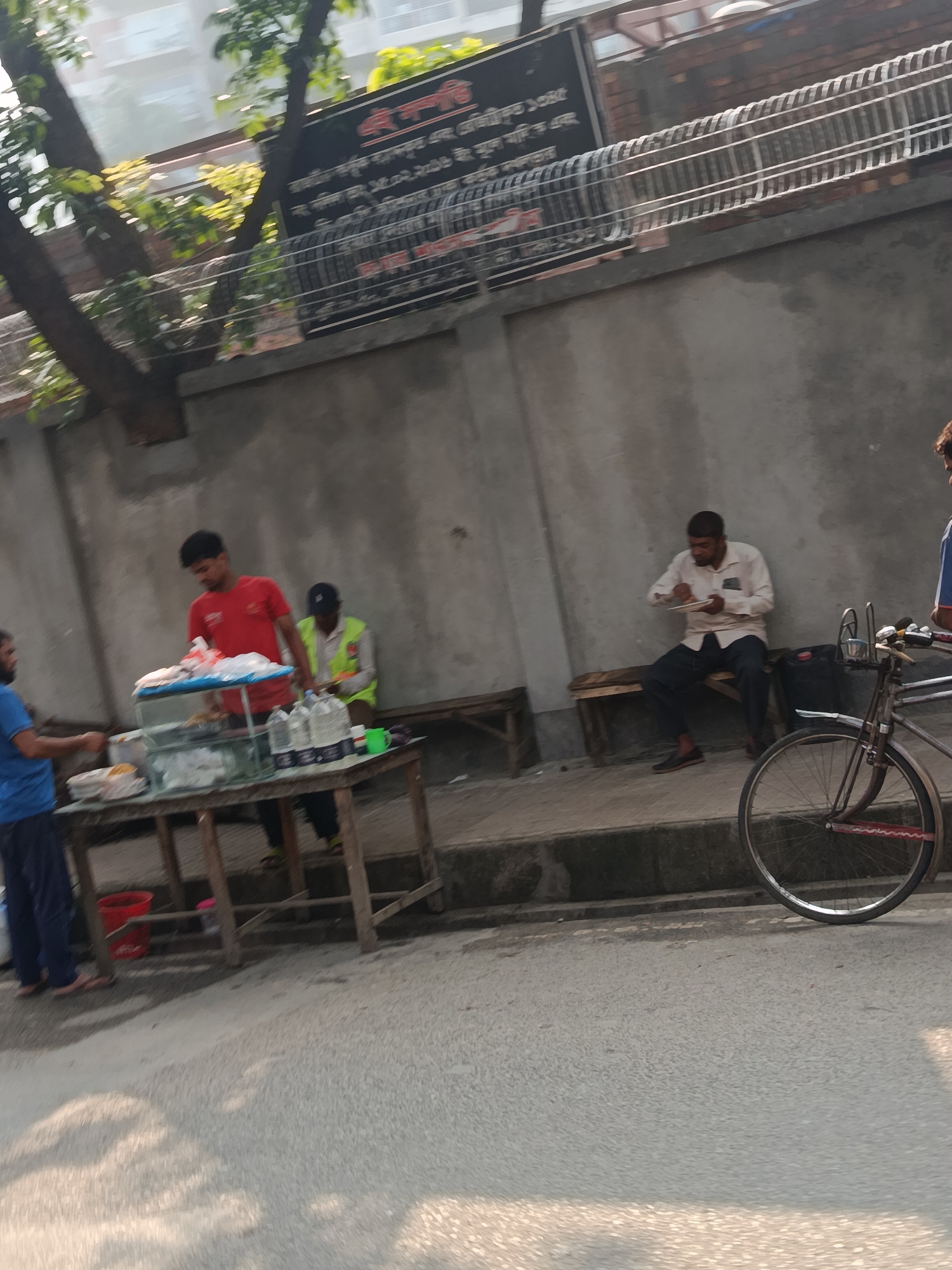 The image depicts a street scene in Dhaka, Bangladesh. A man in a red shirt is selling food from a cart on a sidewalk. Another man, dressed in a blue shirt, stands to the left of the cart appearing to be a customer. A third individual, wearing a neon vest, is sitting on a bench behind the cart. A fourth person is sitting on a bench to the right, eating from a plate, with a bicycle parked in front of him. A concrete wall runs parallel to the sidewalk and there is a sign with text in Bengali above it.