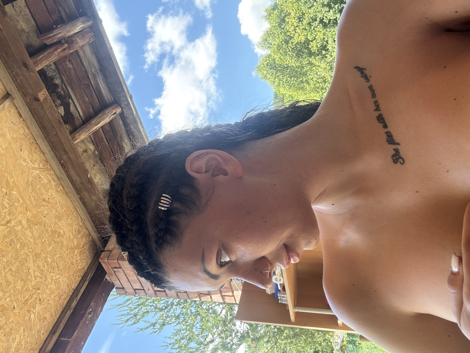 The image depicts a young woman with braided hair, wearing a nose ring and a tattoo on her upper chest that reads "She flies with her own wings." She is positioned outdoors, partially covering her breasts with her hand. The background reveals a wooden structure overhead, possibly a porch roof, with visible beams and insulation. Beyond this structure, lush green trees and a blue sky with white clouds are visible. The setting appears to be a rural or outdoor area, possibly a backyard or a campsite, consistent with a location in Belopoptsi, Bulgaria. The woman is looking off to the side, with her body angled towards the viewer. A shelf unit with bottles is visible to the left of the frame.