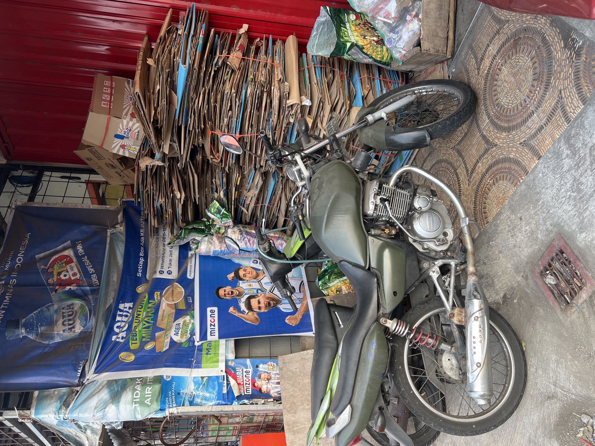 This image depicts an outdoor scene in Tlogoadi, Indonesia, likely in front of a shop or stall. A parked olive-green motorcycle with a silver exhaust pipe is the central focus. In the background, a large stack of flattened cardboard boxes is piled high, some tied with red string, against a red corrugated metal wall. Several large blue and white banners advertising "AQUA" water and "MIZONE" are displayed prominently. One banner features images of football players, including Lionel Messi. To the right of the cardboard stack, a wooden crate contains bags of what appears to be trash or recycling. The ground is a patterned tile, and a metal grate is visible in the foreground. The overall impression is one of an active marketplace or recycling point.