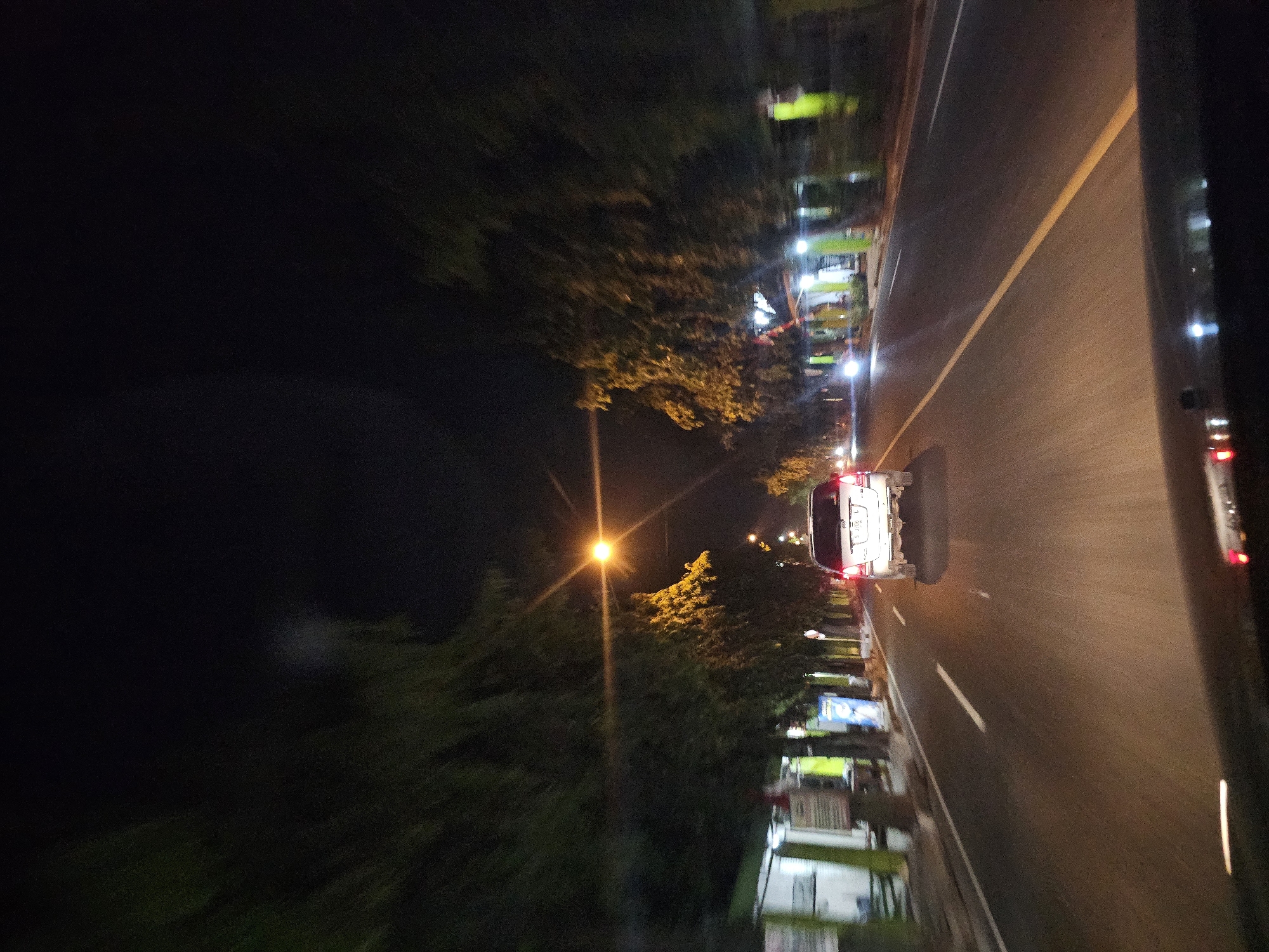 This image depicts a nighttime view from inside a vehicle traveling on a road in Semanding, Indonesia. The road is paved and marked with a white line in the left lane and a yellow line delineating the center of the road. A white SUV is visible ahead, driving in the same direction. The road is lined with trees on both sides, and streetlights illuminate the scene. On the left side of the road, there are buildings or structures with lit windows and some signage. The overall atmosphere is dark, with the primary light sources being the streetlights and the vehicle headlights and taillights. The image appears to be taken from a moving car, as indicated by the slight motion blur, particularly in the trees and the background. There are no people clearly visible in the image.