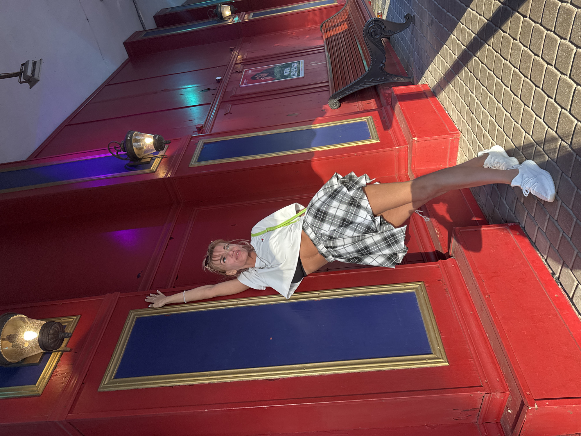 A young woman with blonde hair styled in a messy bun and wearing sunglasses perched on her head poses leaning against a red wall with blue rectangular accents and gold trim. She is dressed in a white crop top, a black bra, and a black and white plaid mini skirt. Her right arm is extended upwards, touching the wall. She is wearing white sneakers. The background features a red wall with decorative lights and a poster. To the right, a wooden bench with ornate black metal legs is visible. The scene appears to be outdoors, possibly on a street or promenade in Saint Petersburg, Russia, indicated by the architecture and the poster text, which seems to be in Cyrillic script. The ground is paved with rectangular stones, and sunlight casts shadows on the ground.