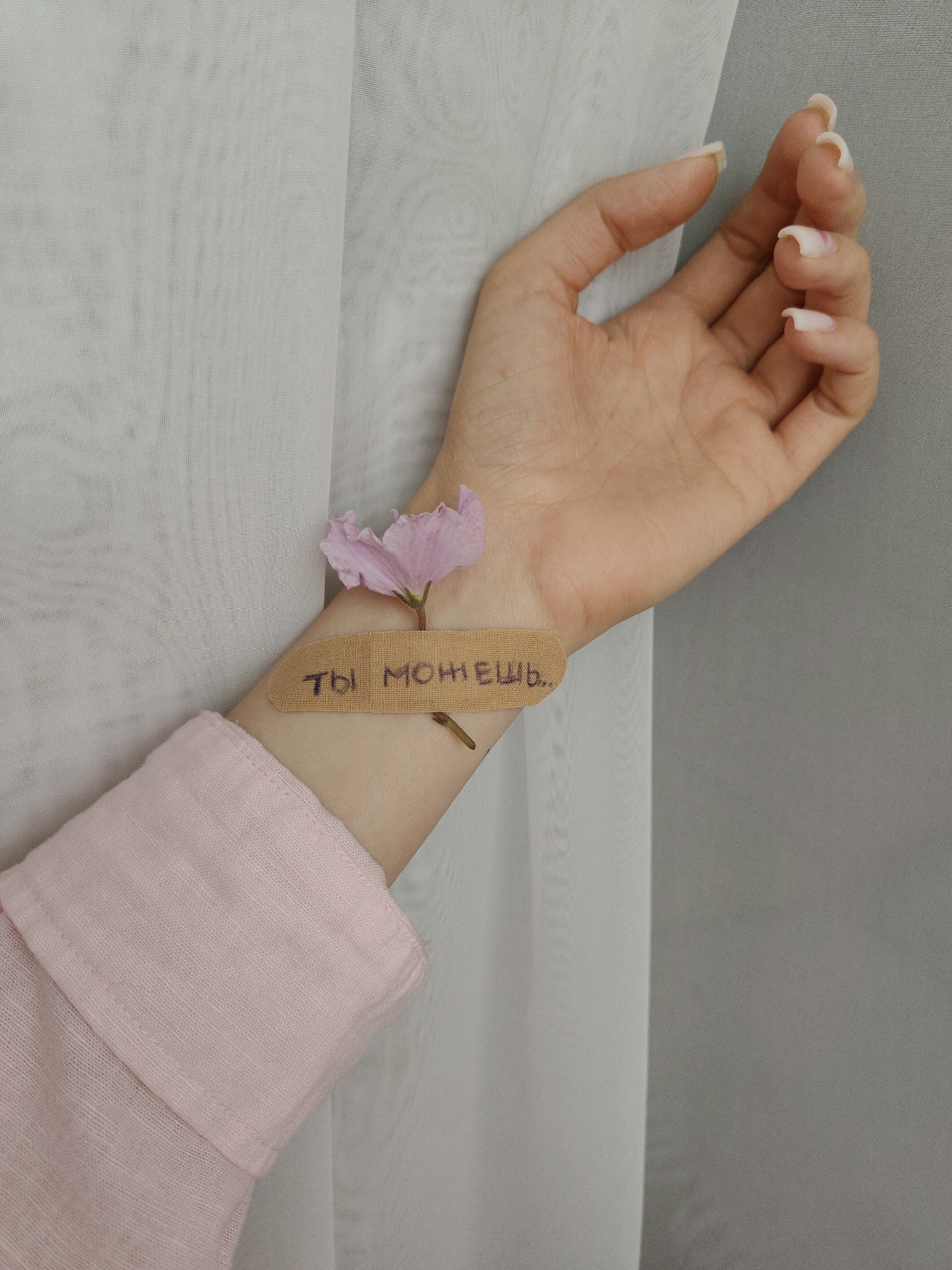 A close-up shot depicts a person's forearm, wrist, and hand. The person wears a pink shirt with the sleeve rolled up. A bandage with the phrase "ты можешь" written on it is placed on the wrist. Atop the bandage is a small, pink flower. The person's hand is extended, fingers slightly curled. The background is composed of white and gray drapery. The location is Unknown, Unknown.