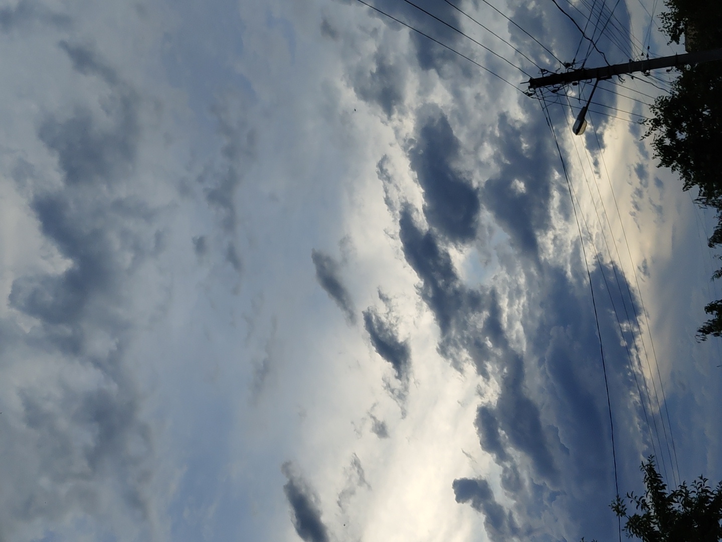 This is a vertical, outdoor, daytime photograph taken from a low angle looking up at a cloudy sky. The sky is filled with dark, heavy clouds, with some lighter patches suggesting the presence of the sun behind them. Several power lines stretch across the frame from the right side. A utility pole with a street lamp attached is visible on the right side of the image, supporting the power lines. Silhouettes of trees are visible at the bottom of the image, suggesting they are in the foreground. The overall impression is a slightly ominous or dramatic sky. The location is Unknown, Unknown.