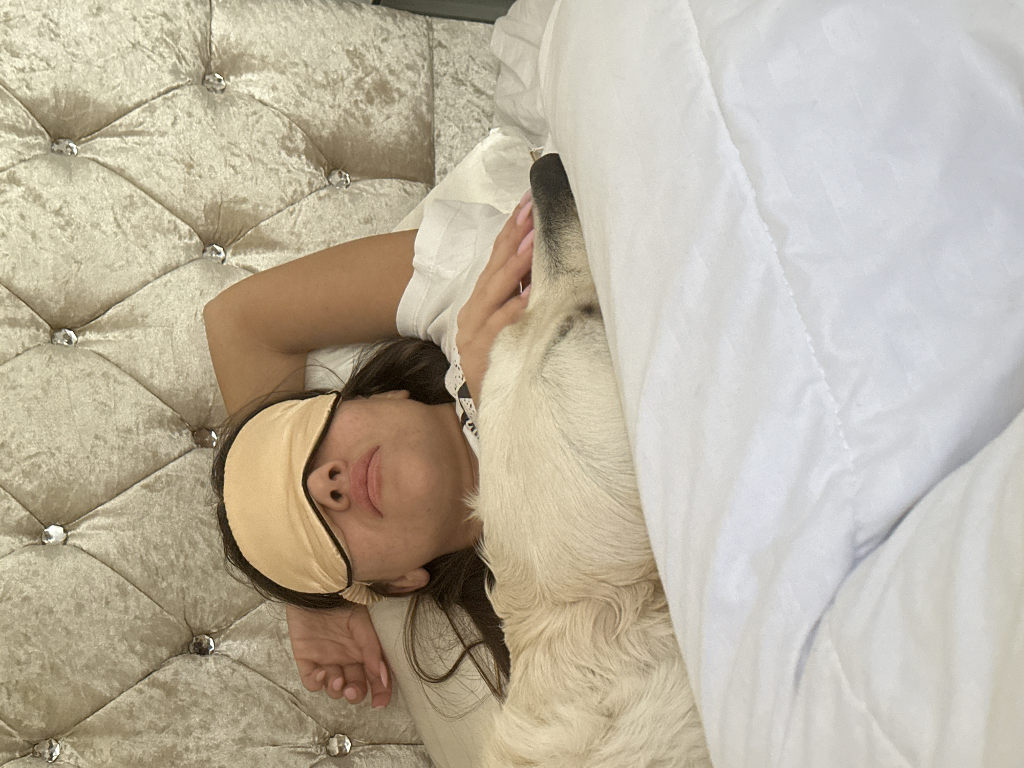 A female subject, appearing to be asleep, lies supine in a bed. She wears a beige sleep mask across her eyes, revealing her nose and lips. Her dark hair is visible around the mask. Her left arm is bent, with her hand resting near the pillow. A light-colored dog, possibly a Golden Retriever, rests beside her, partially obscured by the white quilted bedding. The dog's dark snout and nose are visible near her right shoulder. The headboard behind the subjects is upholstered in a light-colored, tufted fabric, possibly velvet, adorned with clear, button-like decorative crystals. The environment is a bedroom. The location is the city of London, United Kingdom.