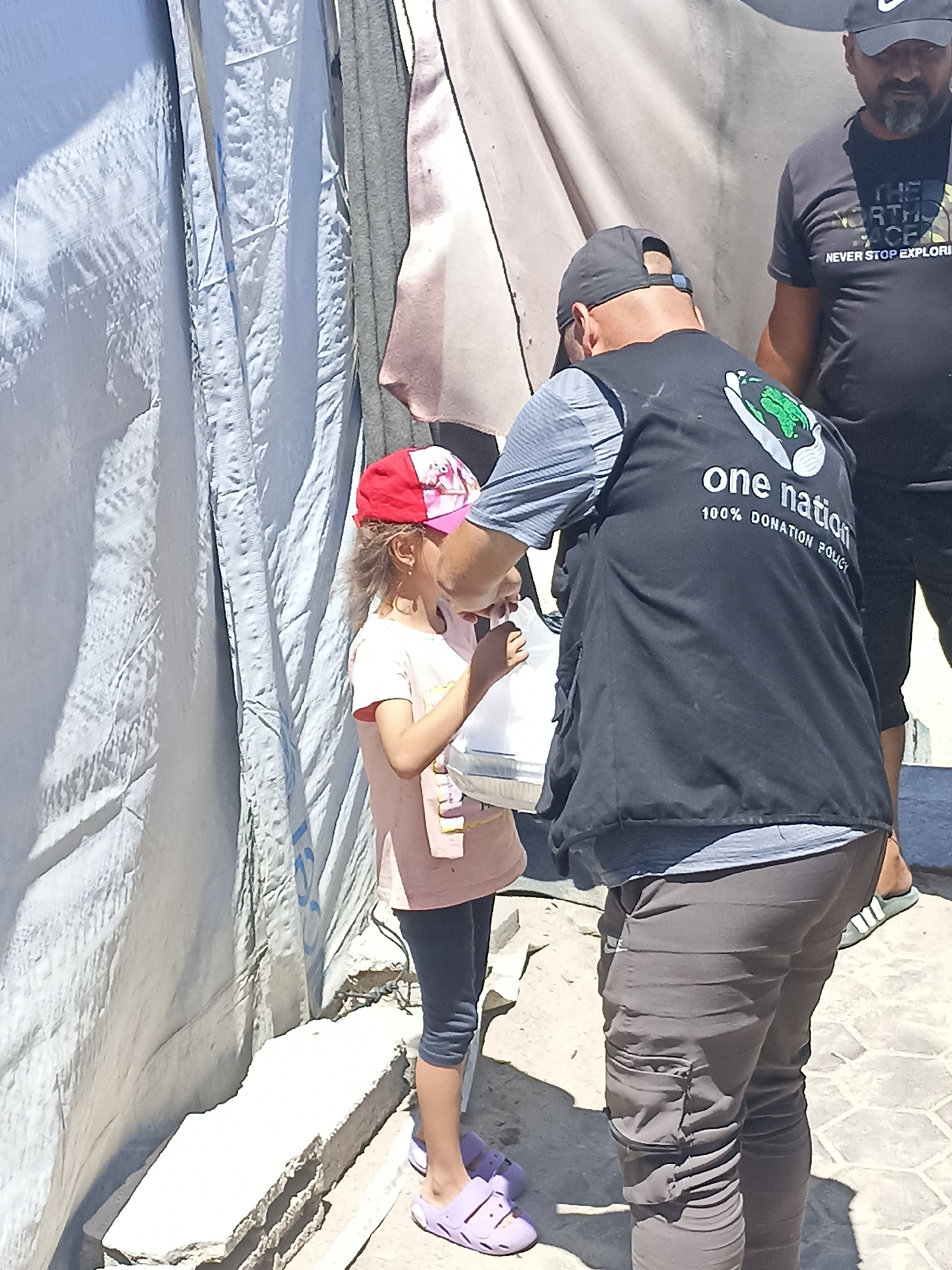 In the city of Gaza, a young girl wearing a pink t-shirt, dark leggings, and a red and pink cap receives an item from a person wearing a black vest with "one nation 100% donation policy" written on the back. The person in the vest is wearing a grey t-shirt underneath and dark pants. Another man wearing a black t-shirt with "NEVER STOP EXPLORING" and a black baseball cap stands in the background. They are in an outdoor area with a large, light-colored tarp or tent in the background and a stone surface below. The scene appears to be a distribution of goods or aid.