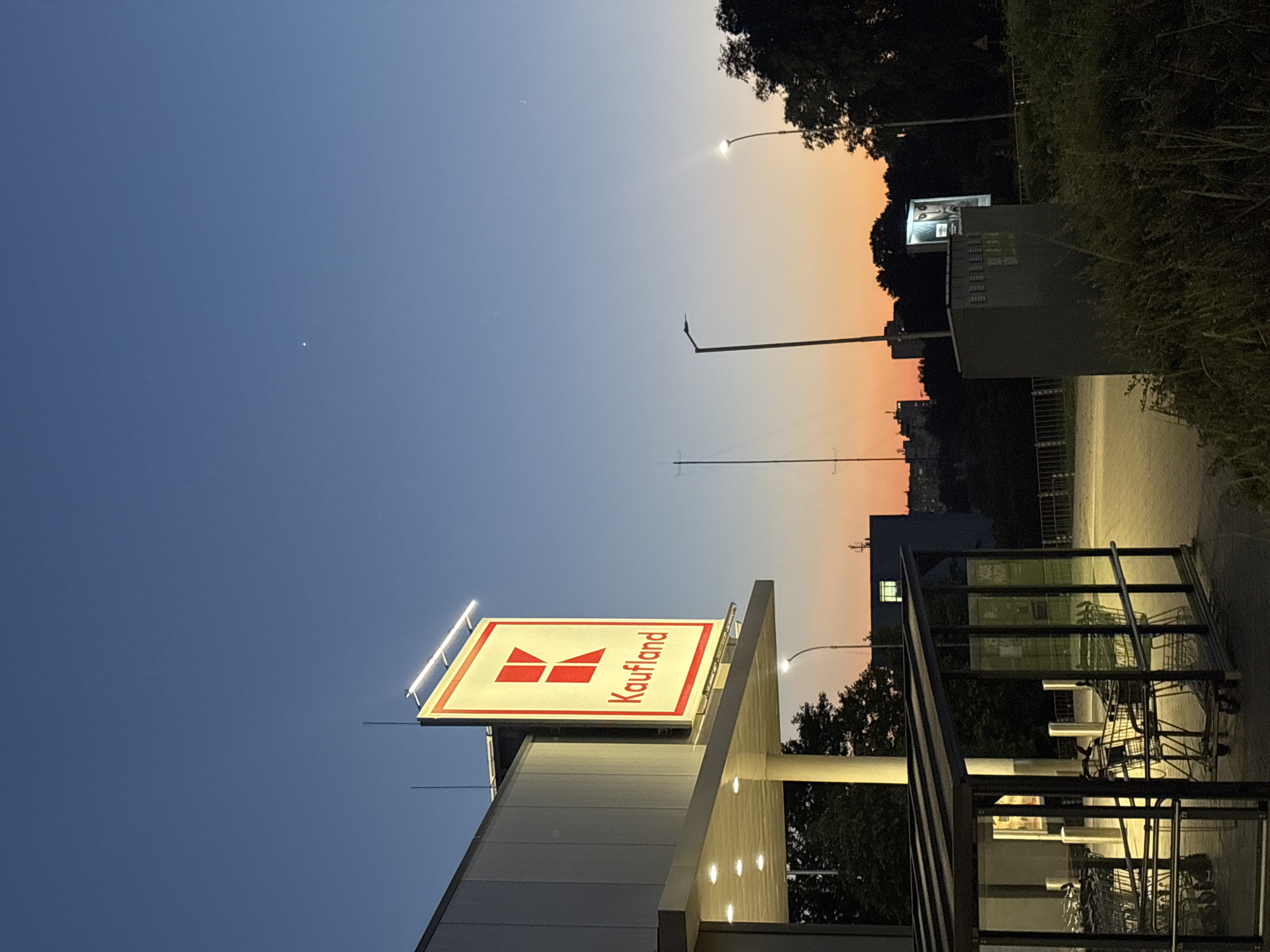 The image shows the entrance of a Kaufland supermarket in Varna, Bulgaria, at dusk. The sky transitions from a deep blue at the top to an orange hue near the horizon. The Kaufland sign, illuminated by lights, is prominently displayed on the left side of the building. In front of the entrance, a covered area with shopping carts is visible, with some carts parked inside. Streetlights are present, and trees and buildings can be seen in the background. The overall atmosphere is calm and quiet, indicating the end of the day.
