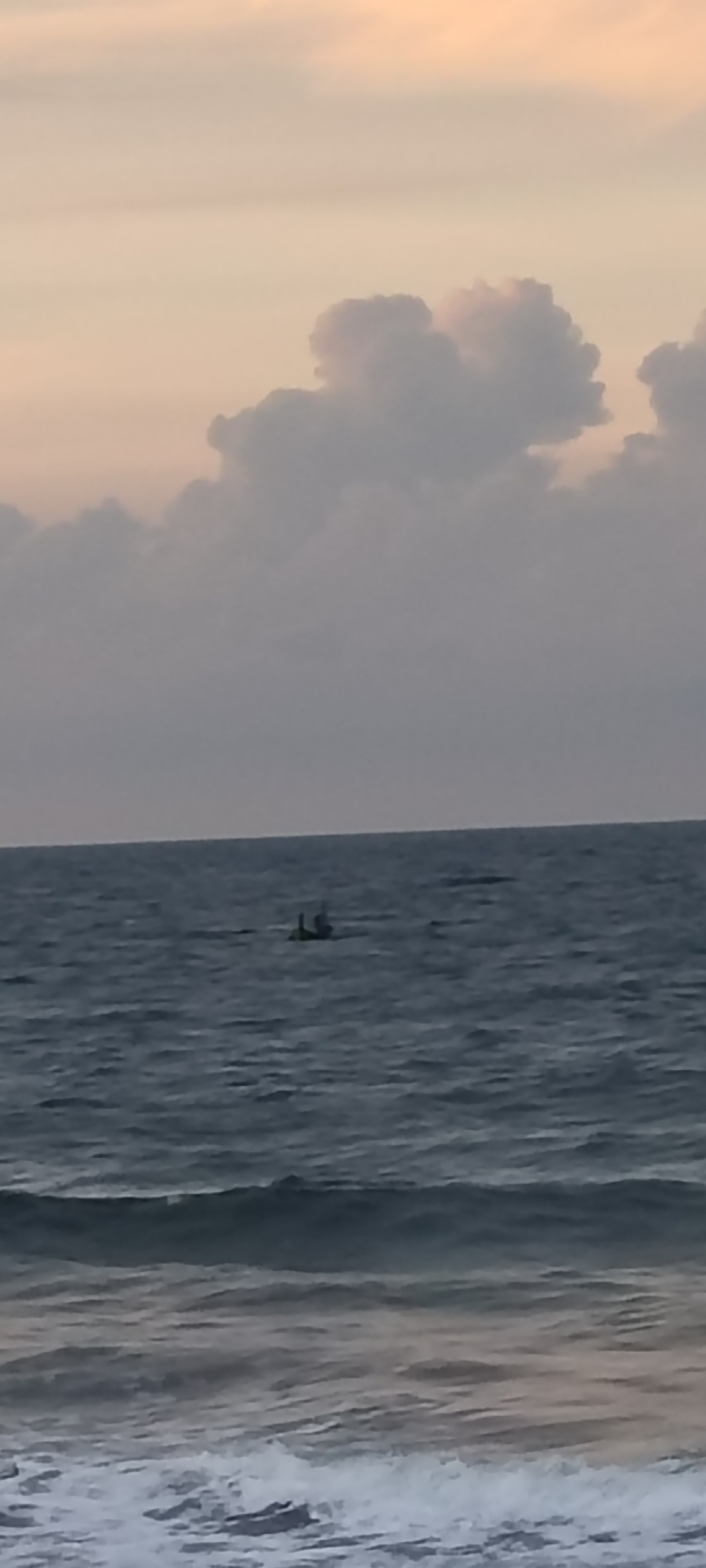 The image shows a view of the ocean at dusk with soft clouds in the sky. A small boat with two people in it is visible in the distance on the water. The waves are gentle, and the overall atmosphere is calm and serene. This scene is likely from the coast of Lhokseumawe, Indonesia, a city known for its beautiful beaches and scenic ocean views.