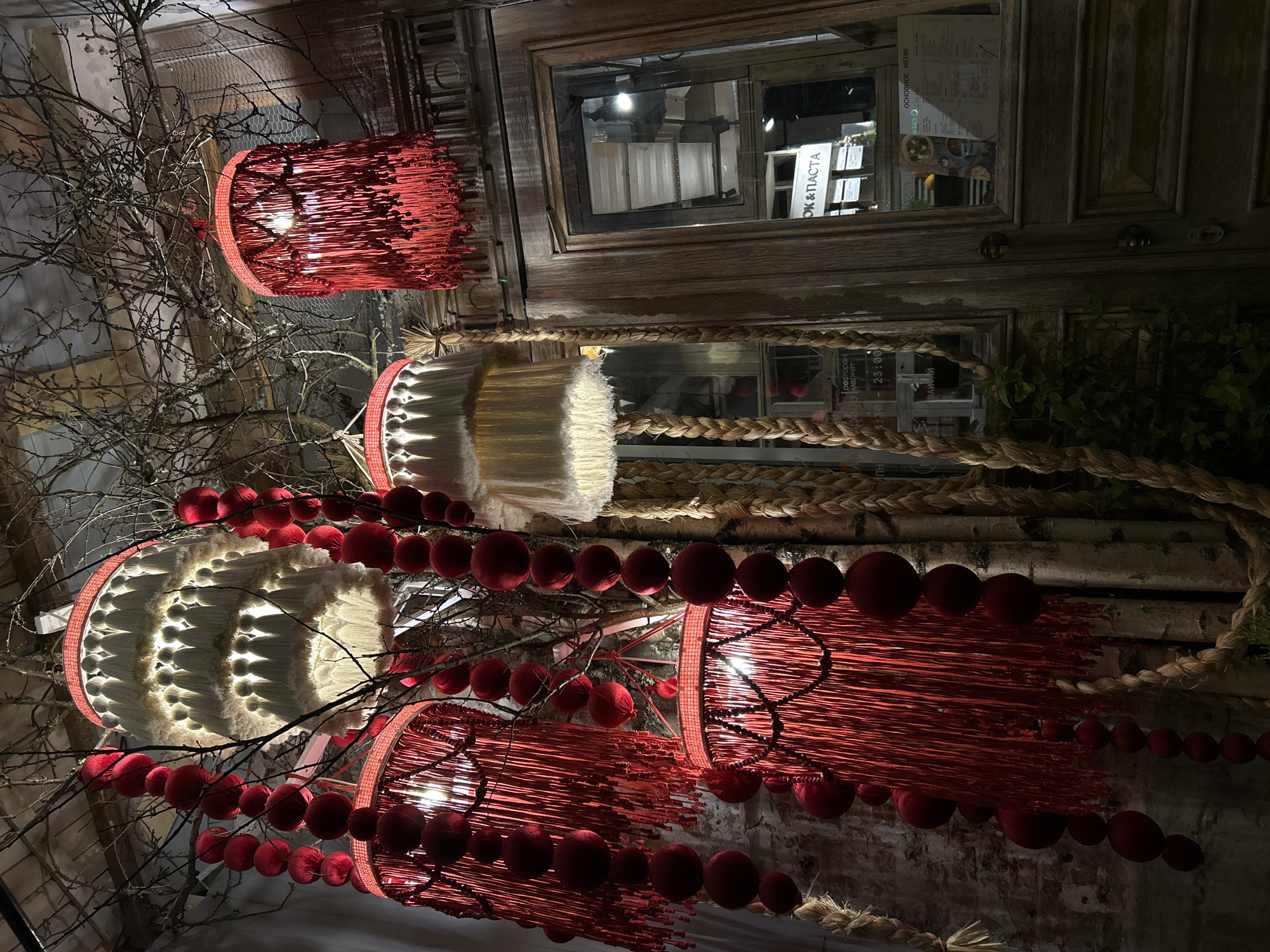 The image depicts a ceiling decoration in Saint Petersburg, Russia. The decorations consist of three hanging light fixtures of differing designs, interspersed with bare tree branches and strings of red, spherical beads. The top fixture is red, cylindrical, and made of thin, vertical strands. The middle and lower fixtures are cylindrical and white, with a row of white, triangular projections along the outer rim. The lower fixture is also red and made of thin, vertical strands.
In the background is a wooden-framed window reflecting the interior of a building. A coarse rope is visible above and to the right of the center fixture. The window frame appears aged and distressed. The surrounding wall is a mix of painted and exposed brick.