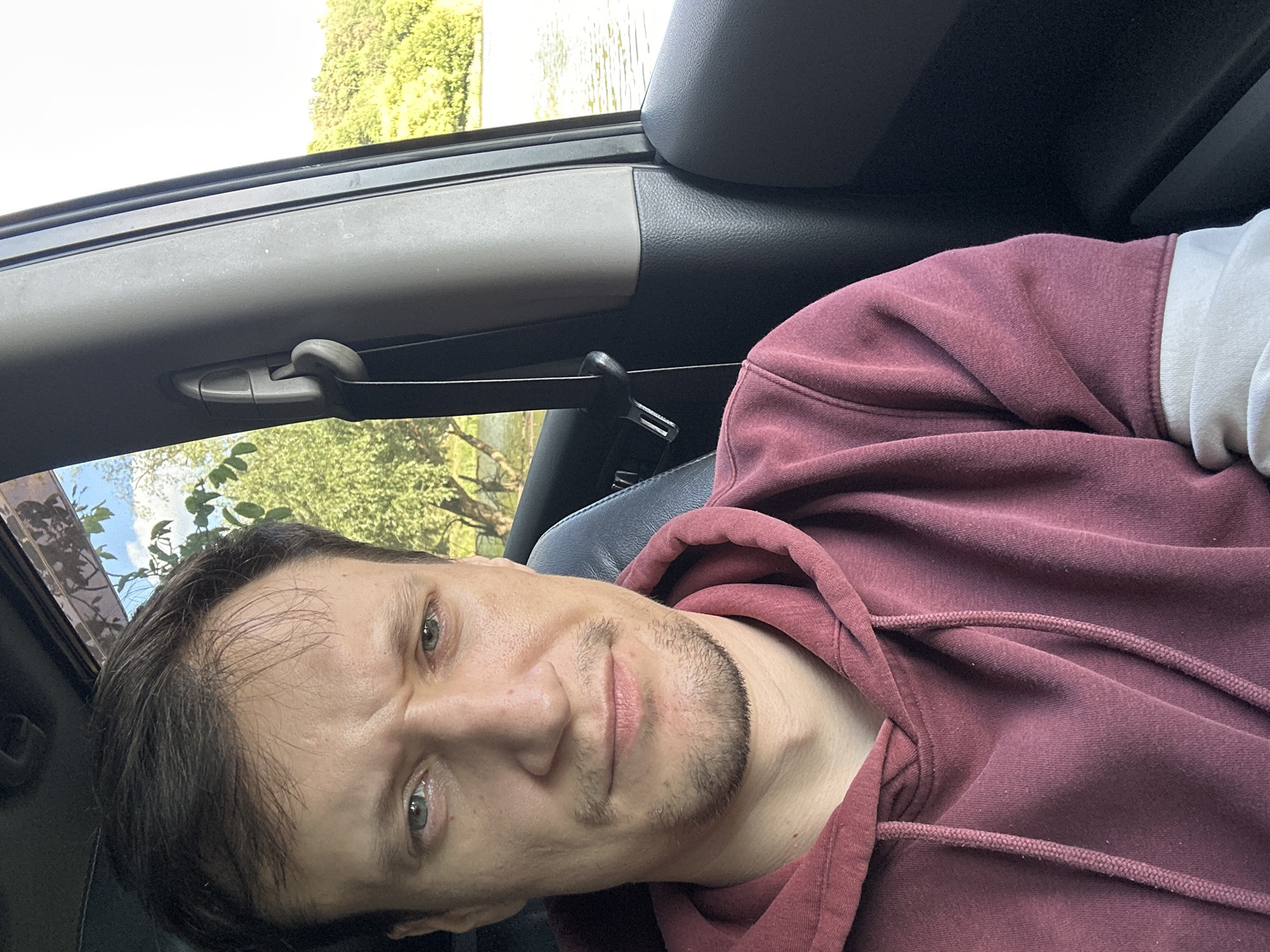 A man is captured in a selfie taken from inside a vehicle, with the scenic backdrop of a lake and lush green trees visible through the car window. He is seated in the driver's seat, looking directly at the camera with a neutral expression. The man is wearing a maroon hoodie with drawstrings. The interior of the car is dimly lit, highlighting the man's face and upper body, while the exterior scene is bright and sunlit. The overall impression is of a casual, candid moment during a drive. The location is identified as Makiv, Ukraine.