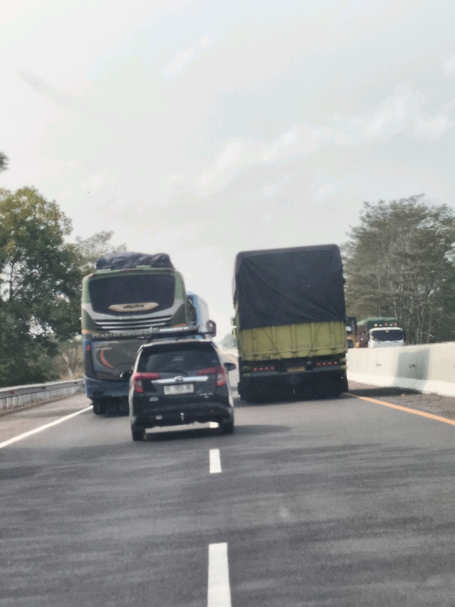 The image captures a highway scene, likely in Lubuk Dalam, Indonesia, given the context. In the foreground, a black car is being filmed from behind. Behind it, a large green and white bus is visible, also seen from the rear, with luggage on its roof. To the right of the bus, a yellow and black truck with a covered cargo bed is moving forward. Further in the distance, other vehicles, including a white truck, are present. The road is paved, with clear white lane markings. Trees and foliage line the sides of the highway, and the sky is overcast with a pale, diffused light. A concrete barrier separates the road from the environment on the right side.